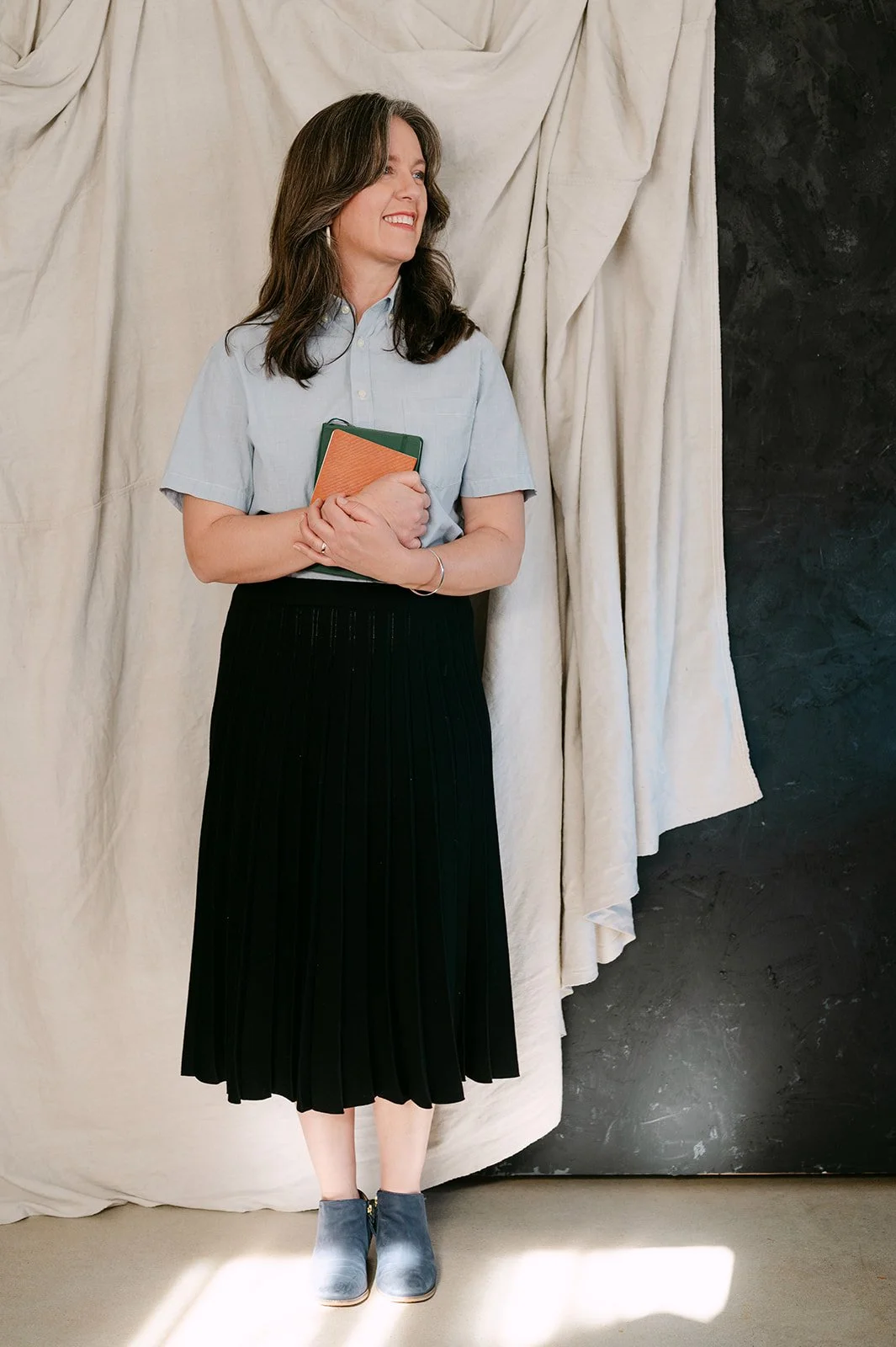 A woman with shoulder-length brown hair, wearing a light blue button-up shirt, black pleated skirt, and gray ankle boots, holding notebooks, standing in front of a cream-colored curtain and a black wall, with a small patch of sunlight on the floor.