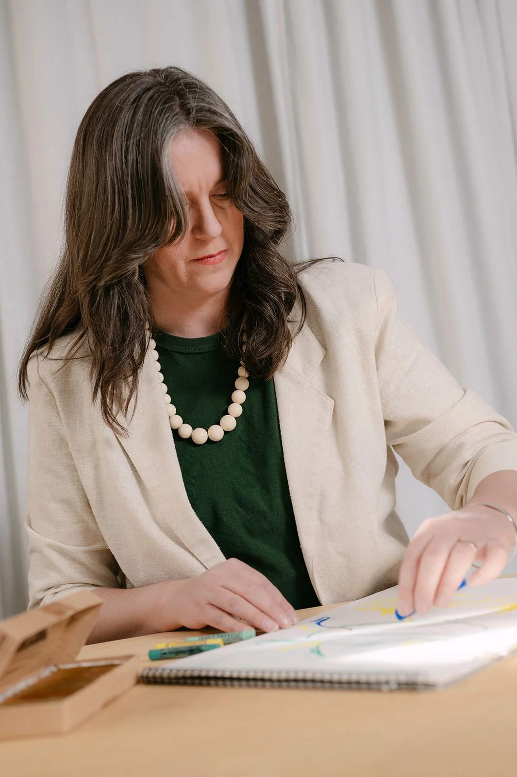 A woman with dark wavy hair, wearing a beige blazer, green shirt, and pearl necklace, is sitting at a desk, drawing or coloring on a notebook with colorful markers.