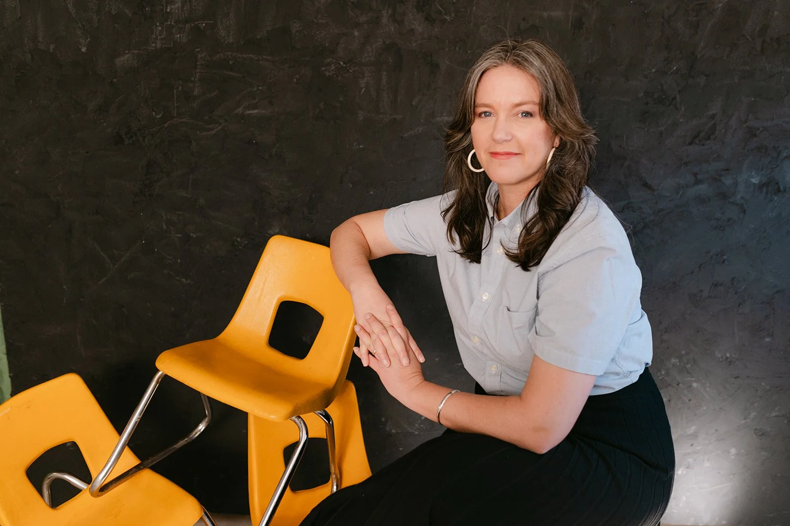 A woman with shoulder-length brown hair wearing a light grey short-sleeve shirt, sitting next to yellow chairs with black cutout backrests, against a dark textured background.