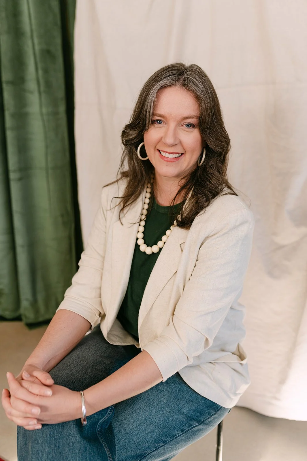 A woman with wavy brown hair in a white blazer, green shirt, and jeans, smiling and sitting against a backdrop with green and white drapes.