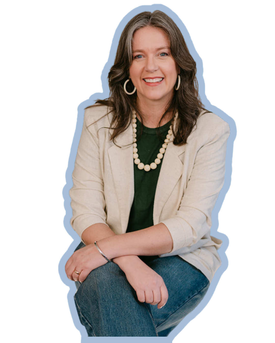 A woman with long brown hair, wearing a cream blazer, green shirt, pearl necklace, hoop earrings, and jeans, smiling with her arms crossed in a seated pose.