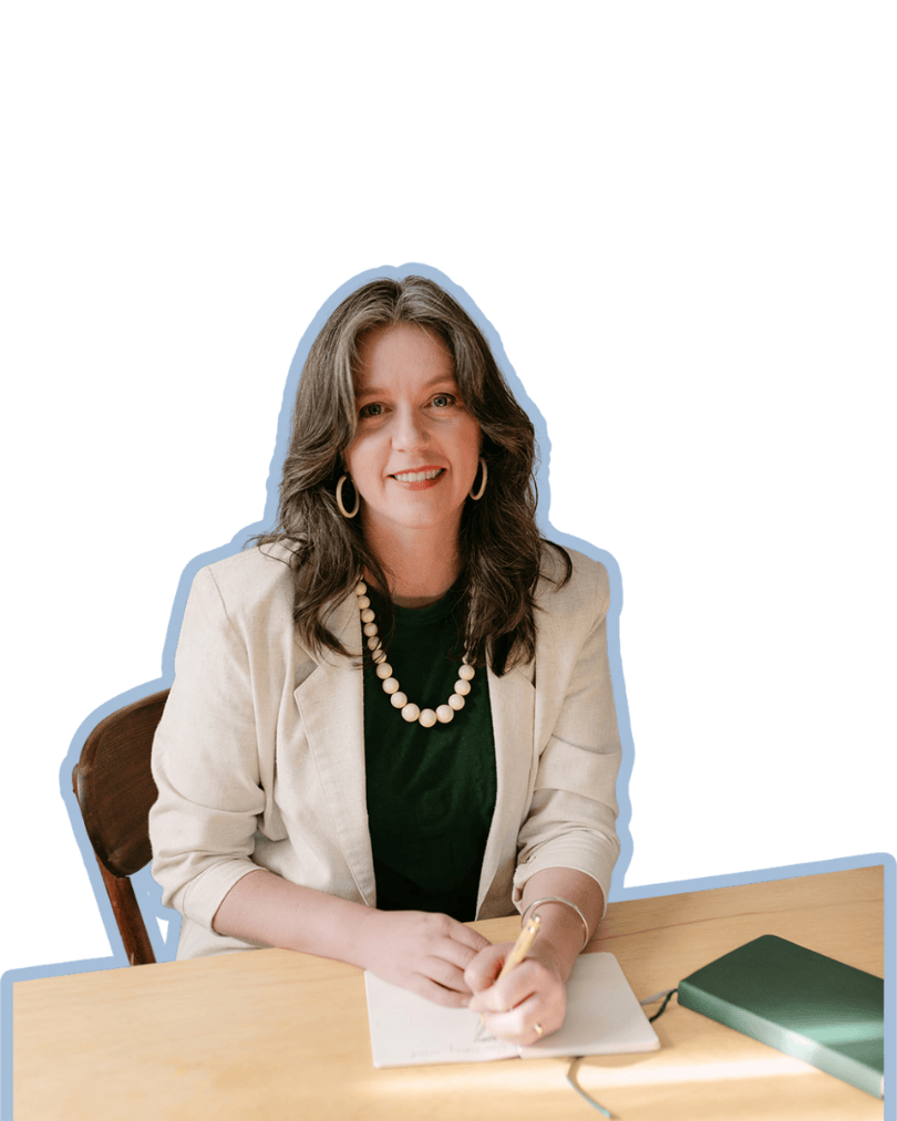 A woman with shoulder-length dark hair, wearing a cream blazer, black top, pearl necklace, and hoop earrings, sitting at a light wooden desk with a notebook and green book, smiling at the camera.
