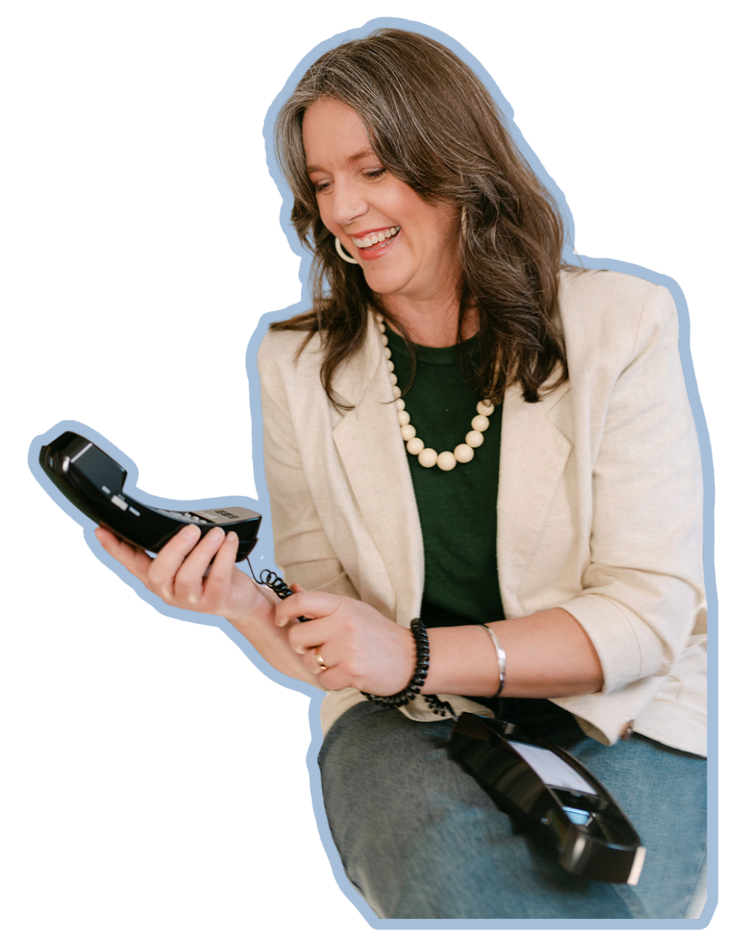 A woman with brown shoulder-length hair, wearing a beige blazer, green shirt, a pearl necklace, and bracelets, smiling while holding a landline phone.