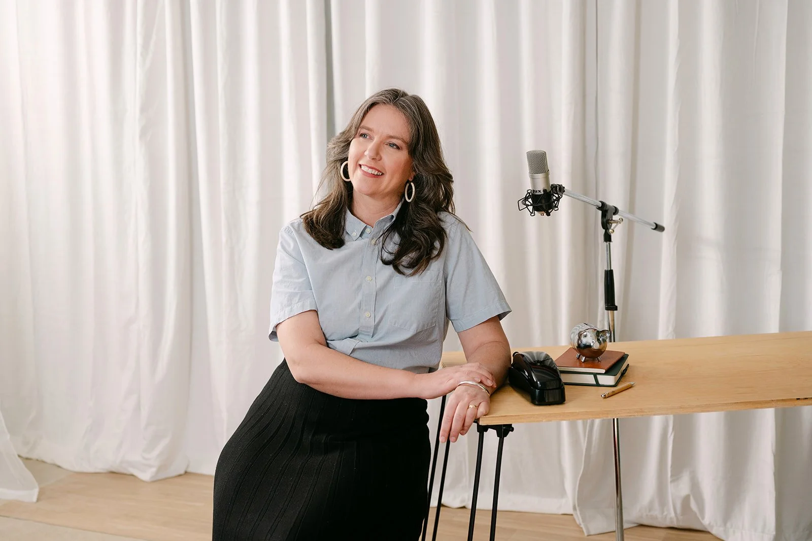 A woman with dark wavy hair, wearing a light blue button-up shirt and a black skirt, standing beside a wooden table with a microphone, notebook, pen, and small silver piggy bank, in front of white curtains.