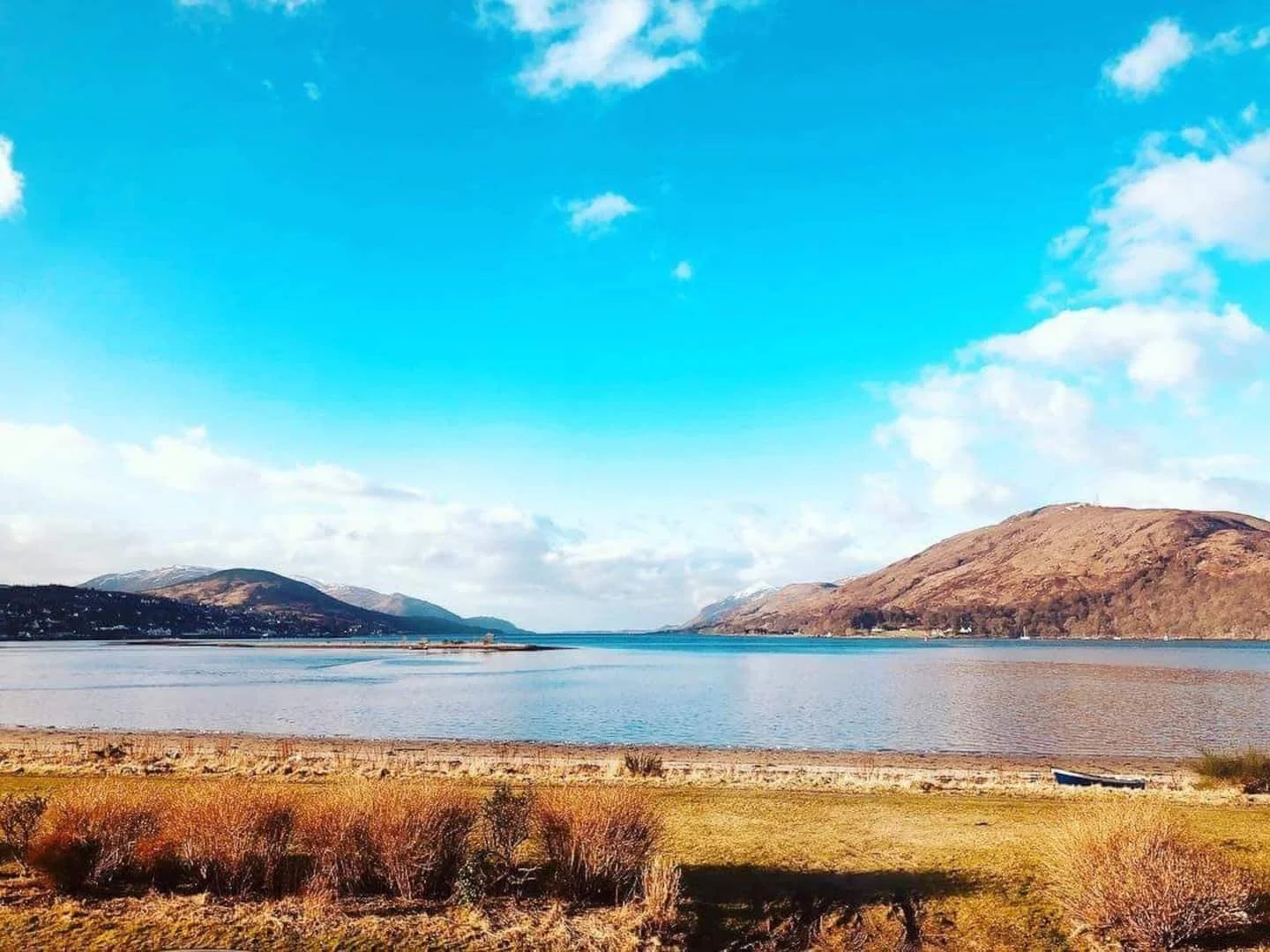 A scenic landscape of a lake with hills and mountains in the background, blue sky with scattered clouds, and dry vegetation in the foreground.