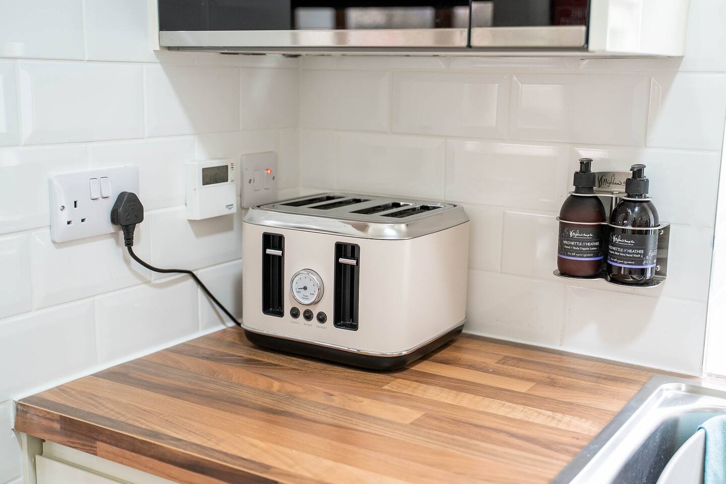 Toaster on a kitchen countertop next to a wall outlet with a plugged-in black cord, and a shelf with two brown bottles labeled moisturizer and lotion.