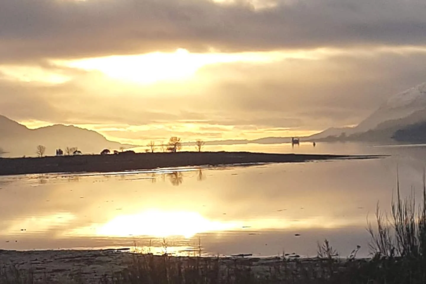 Sunset over a body of water with mountains in the background, cloudy sky, and some trees and plants near the shore.