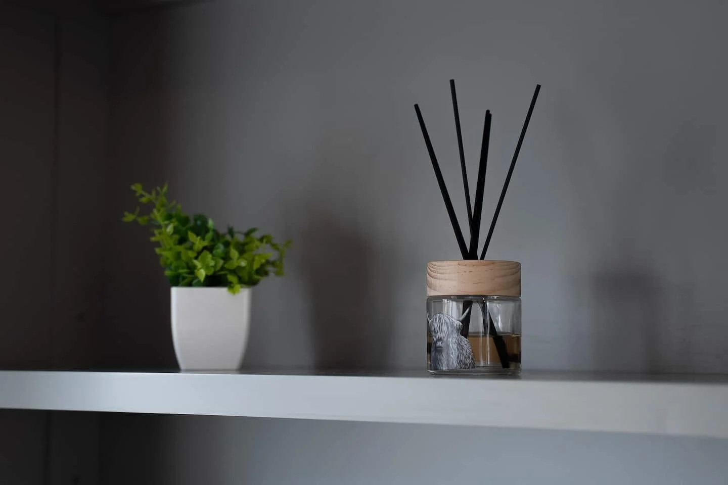 A white shelf with a small green potted plant on the left and a glass reed diffuser with black sticks and a wooden lid on the right.