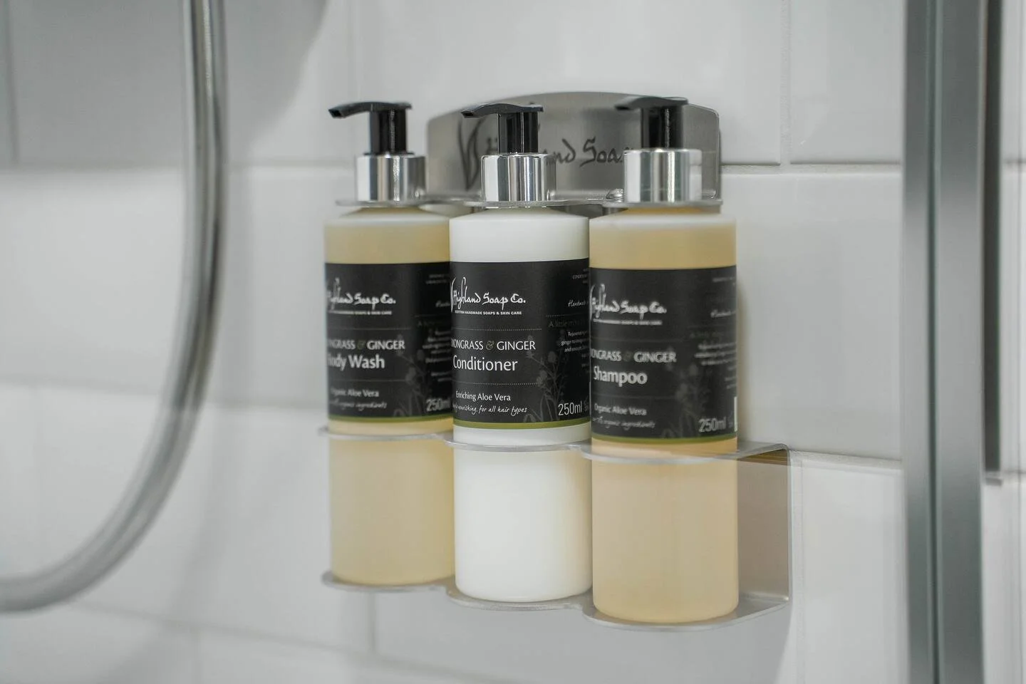 Three bottles of Highland Soap Co. shampoo, conditioner, and body wash on a bathroom shelf, with the labels indicating ginger and aloe vera ingredients, in a shower area.
