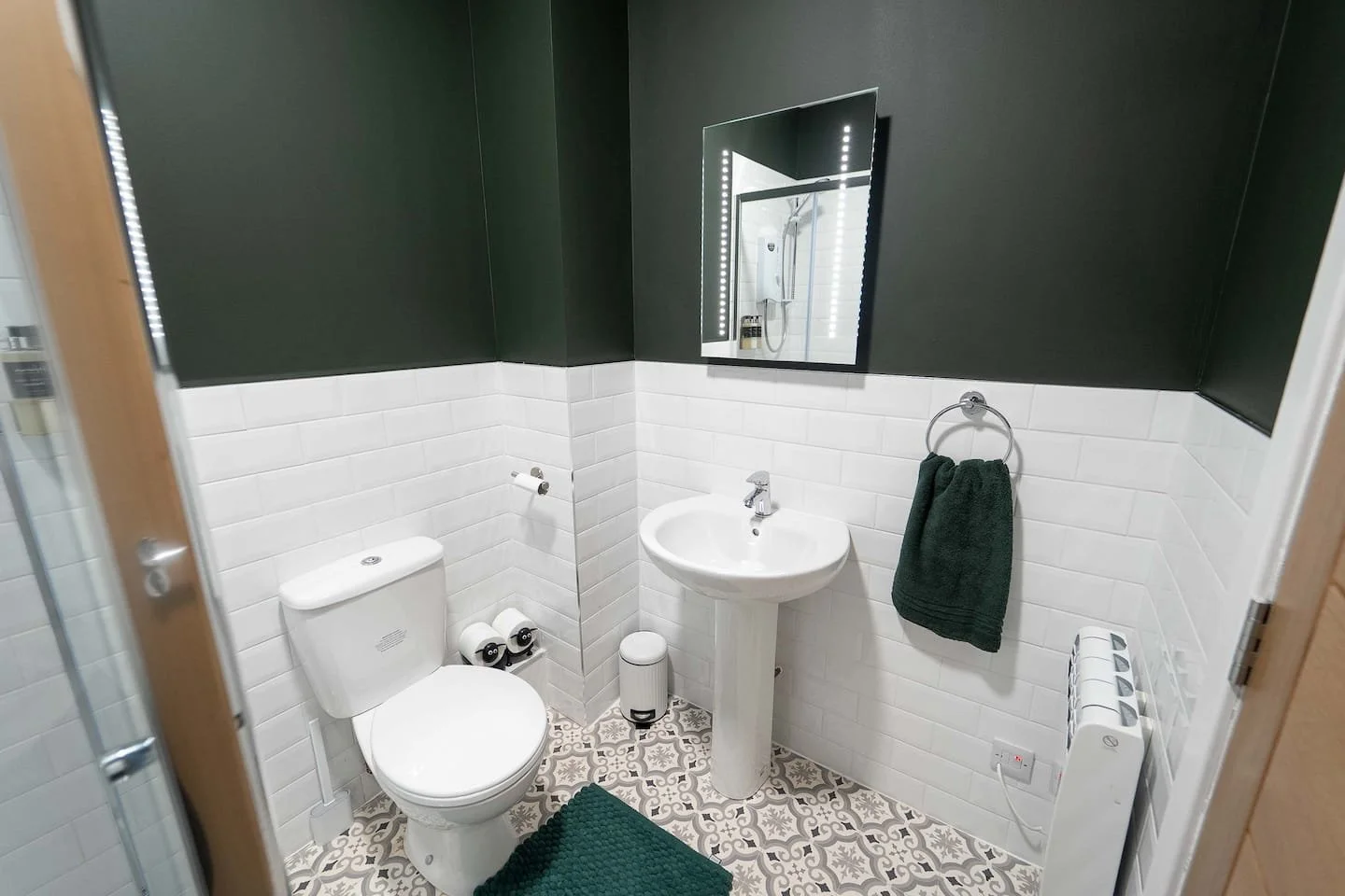 Small modern bathroom with white subway tile walls, a toilet, a pedestal sink, a mirror with LED lights, a dark green towel on a ring, a patterned tile floor, and a small trash can.