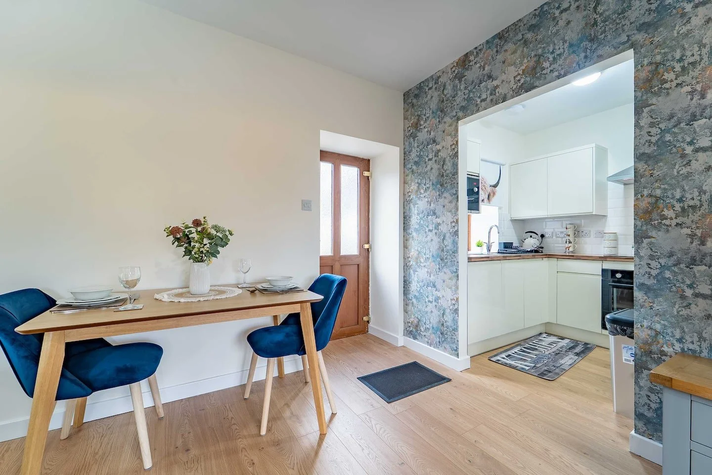 Dining area with a wooden table set with white plates, glasses, and a vase of flowers, adjacent to a kitchen with white cabinets and a textured wall.