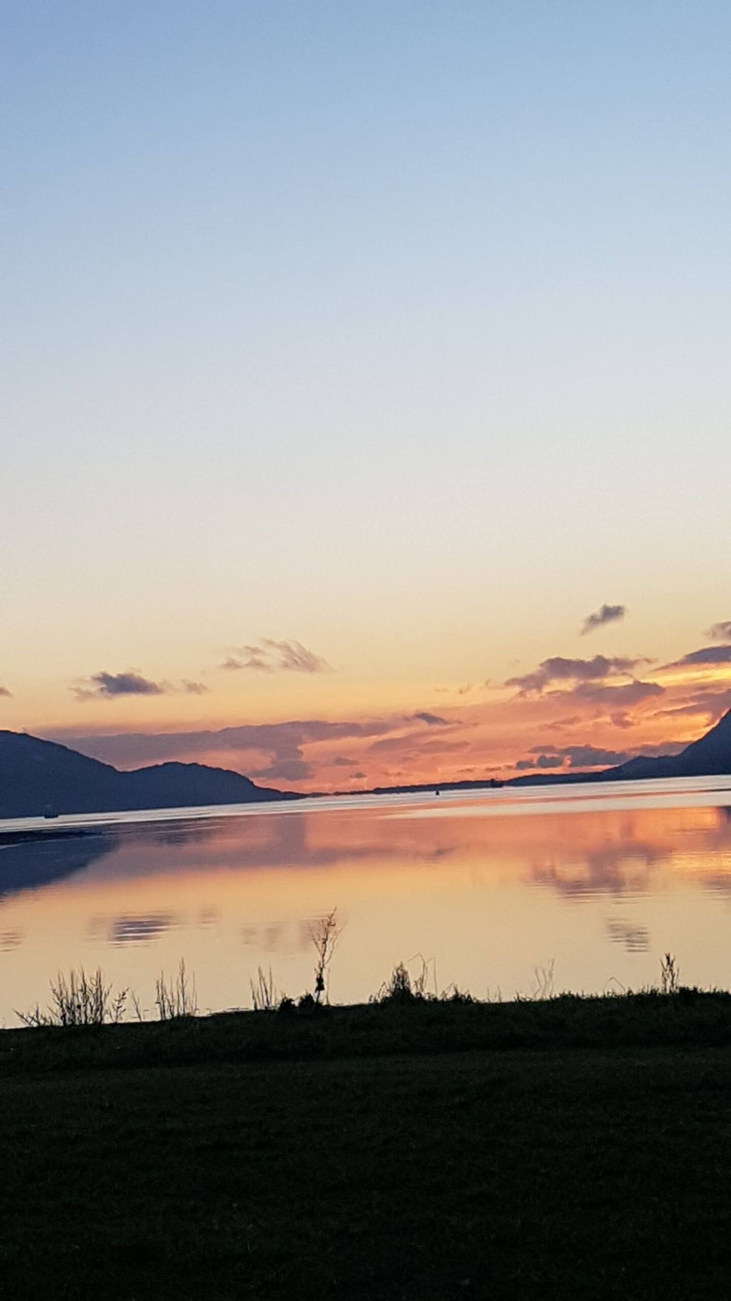 Serene lake at sunset with mountains in the background, calm water reflecting the colorful sky, some small trees and bushes in the foreground.