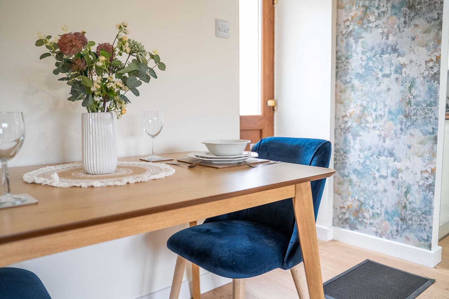 A dining table set with white bowls, plates, wine glasses, a vase with flowers, and a lace doily, with a blue velvet chair nearby and a blue textured wall in the background.