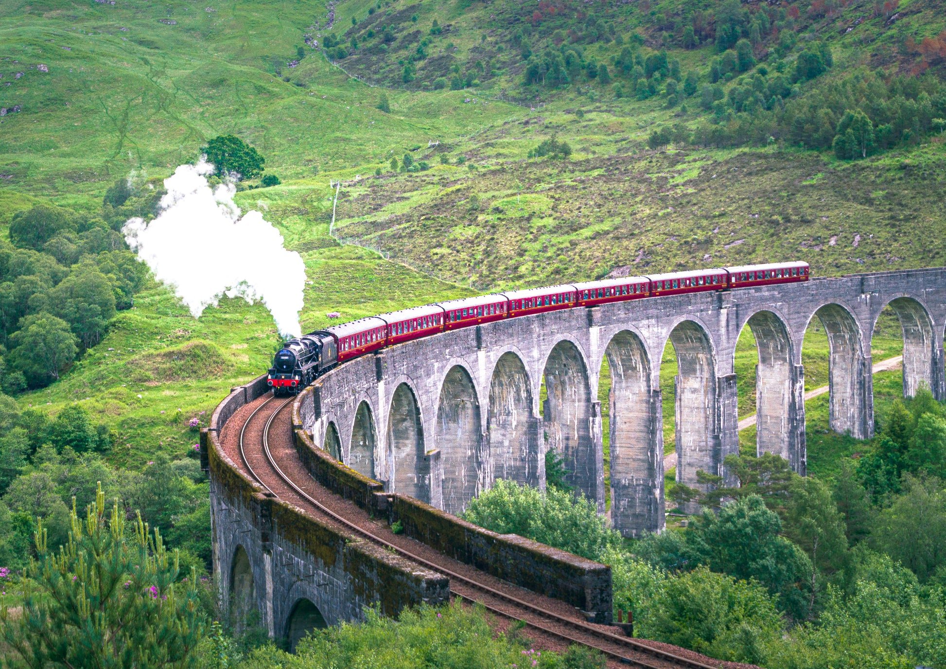 A steam train with red cars traveling over a large stone viaduct in a lush, green, mountainous landscape.