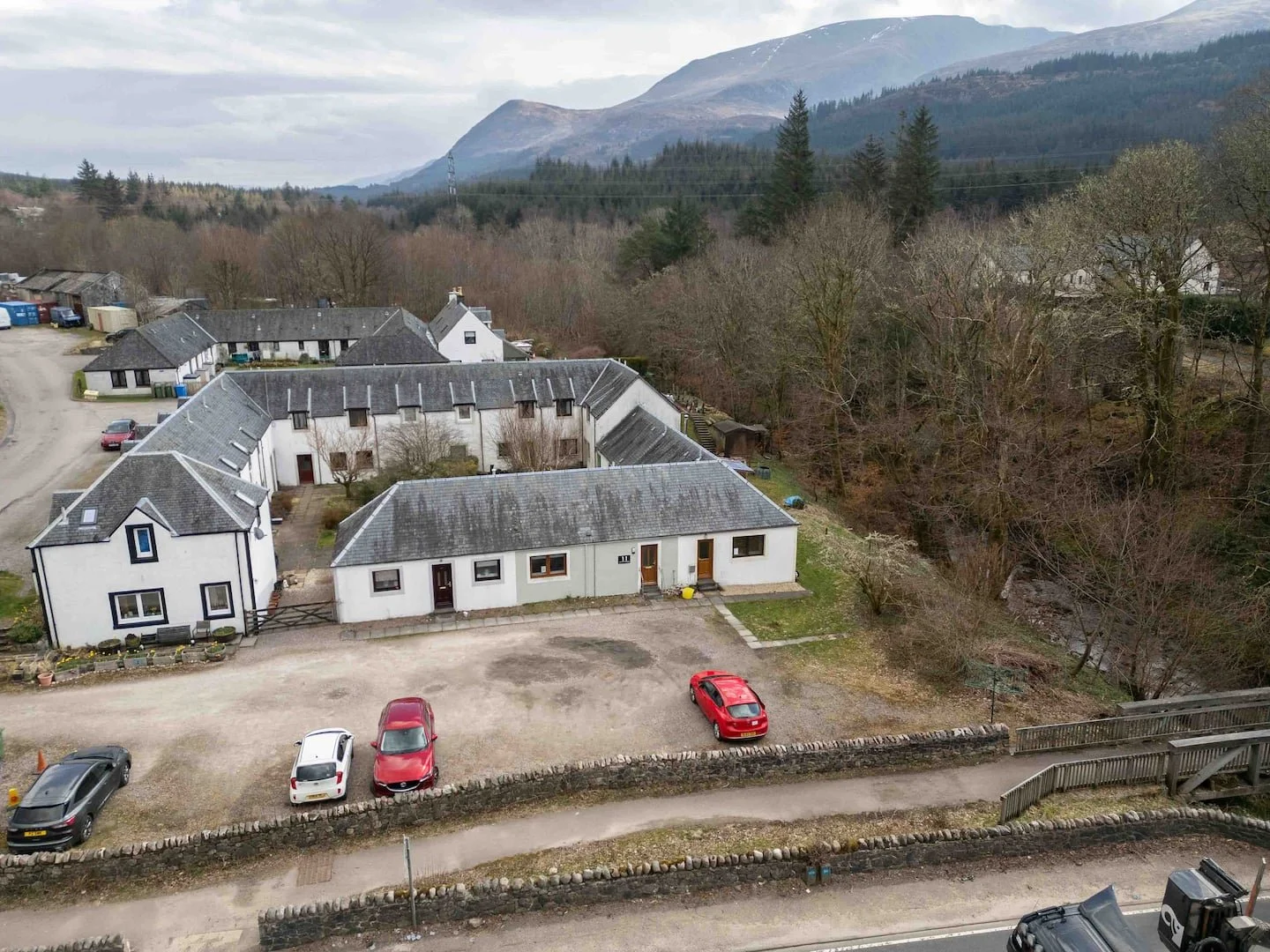 A view of a residential area with white houses, parked cars, surrounded by trees and mountains in the background.
