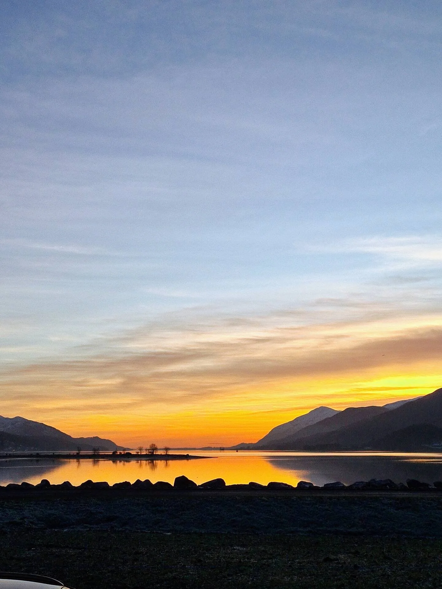 Sunset over a calm body of water with mountains on both sides and a rocky shoreline in the foreground.