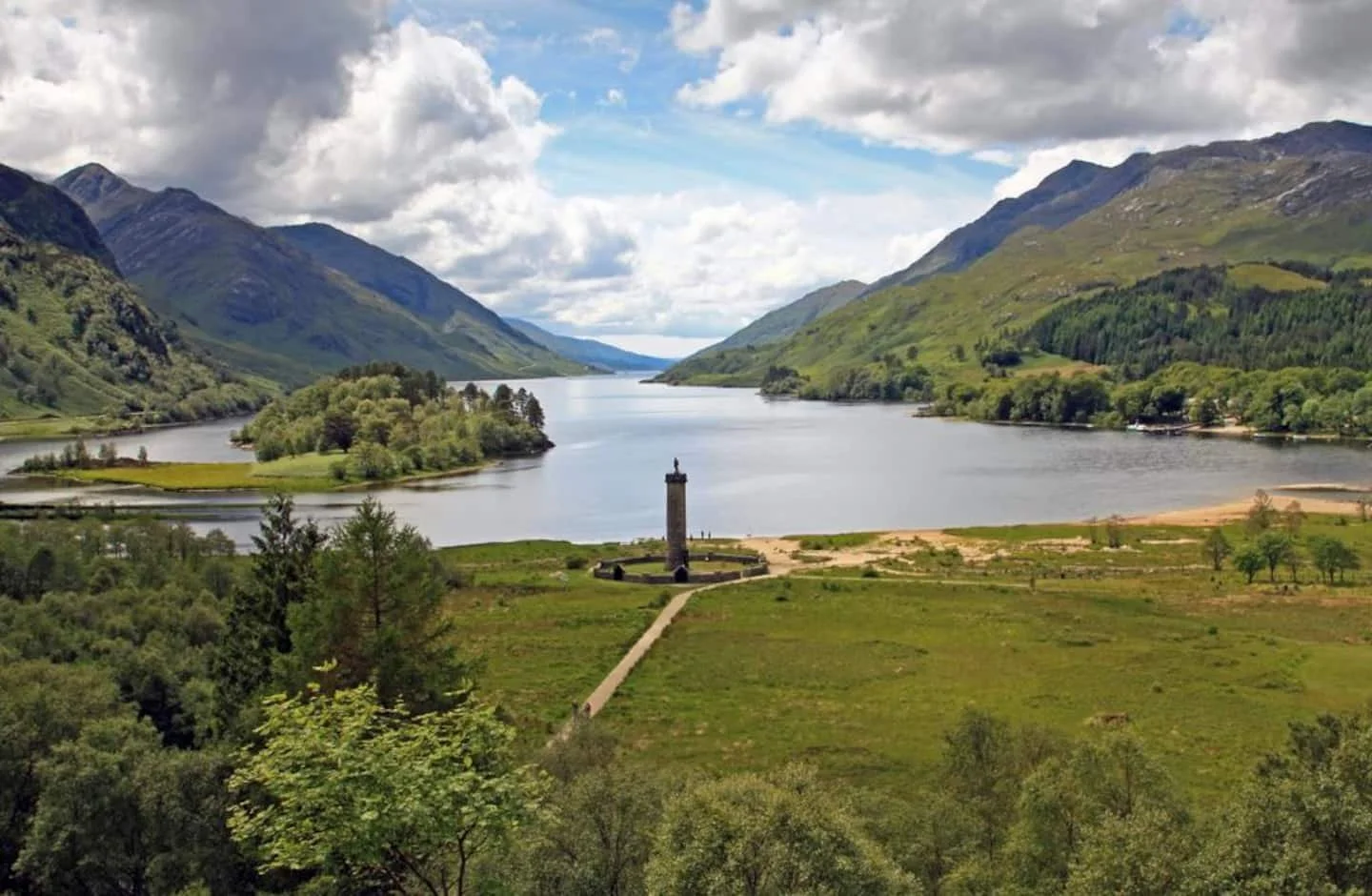 Scenic view of a river or lake surrounded by lush green hills and mountains under a partly cloudy sky, with a tall monument or tower in the foreground.