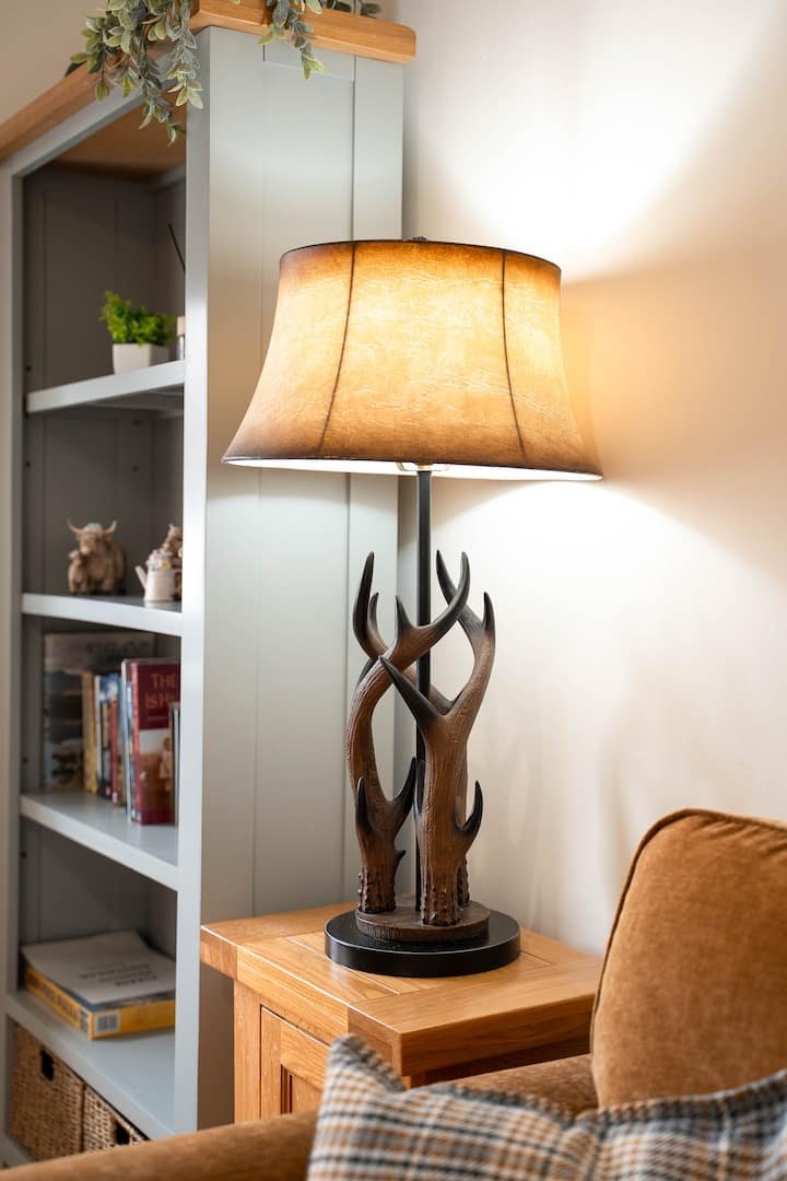 A lamp with a base made of intertwined wooden antlers and a beige lampshade on a wooden side table in a cozy living room corner.