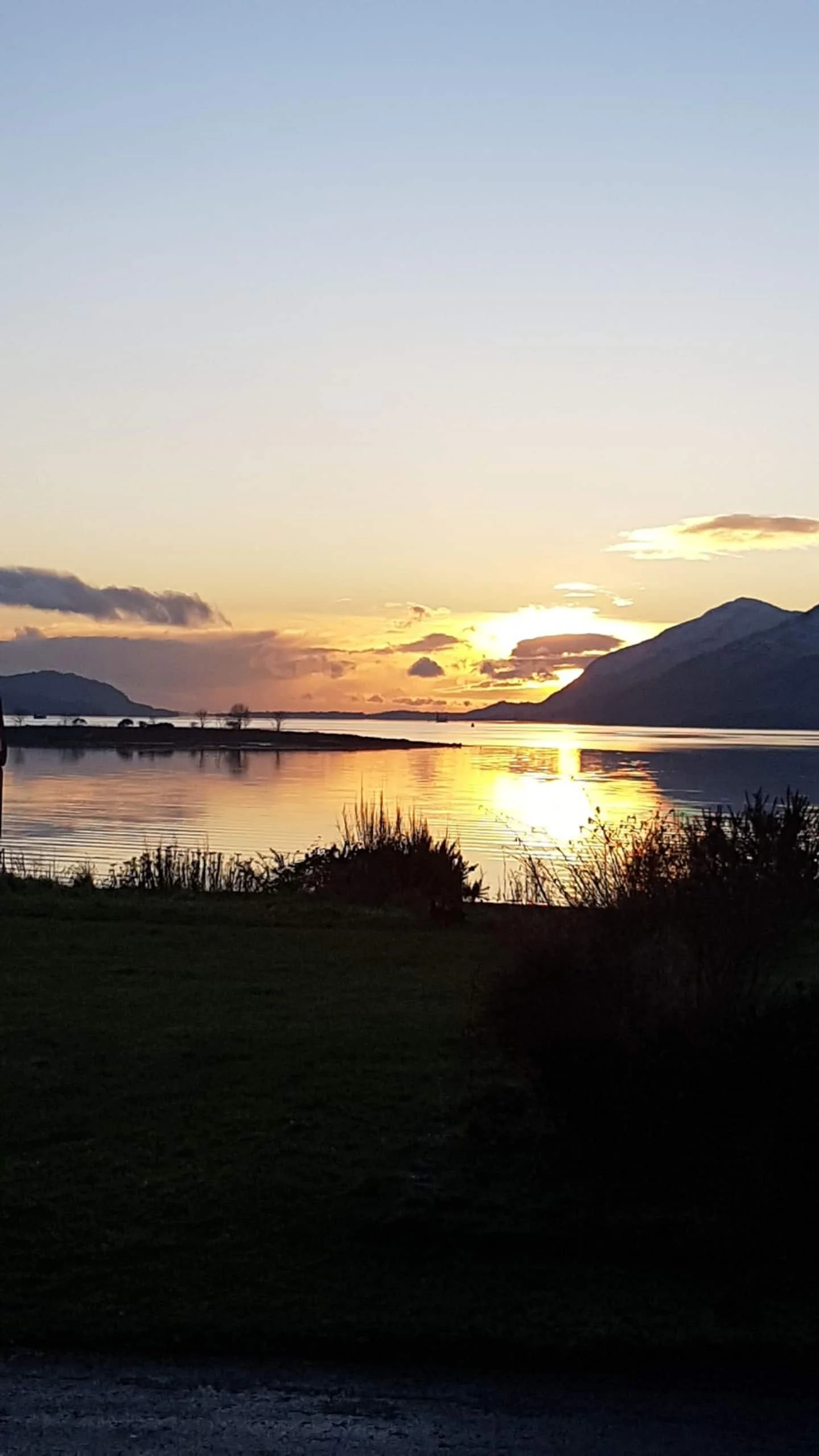 Sunset over a calm lake; mountains in the background; bushes and grass in the foreground.