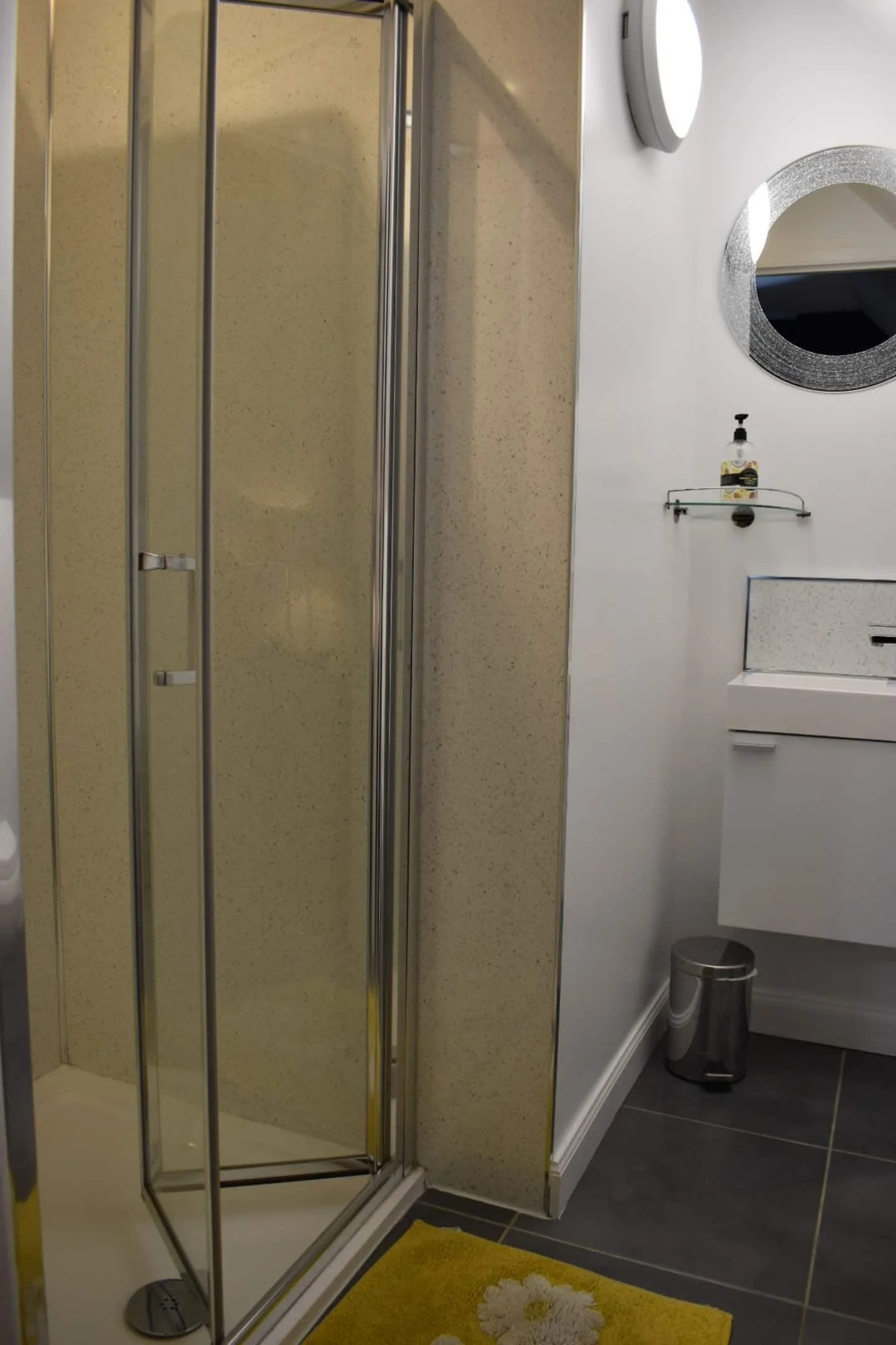 A bathroom corner with a glass shower enclosure, a white sink cabinet, a round mirror with a silver frame, a small shelf with a soap dispenser, a round wall light, a soap pump, a small trash bin, a yellow rug with white floral pattern, and gray tiled