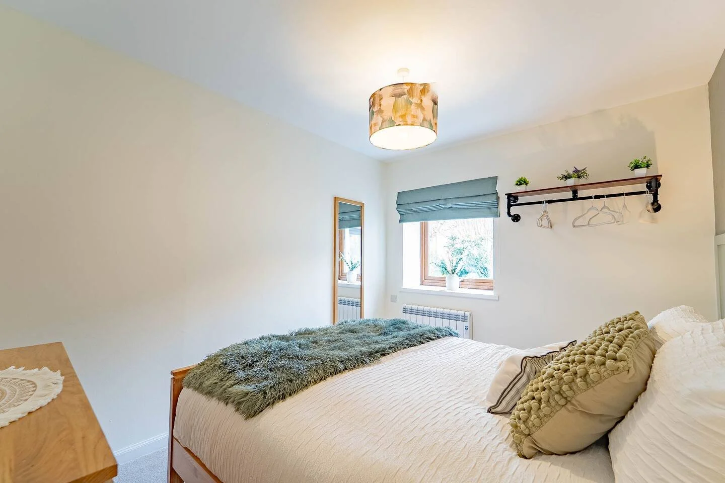 A bright bedroom with a bed, decorative pillows, and a green throw blanket. A window with a blue Roman shade, a mirror, and a wall-mounted shelf with plants and hangers are visible.