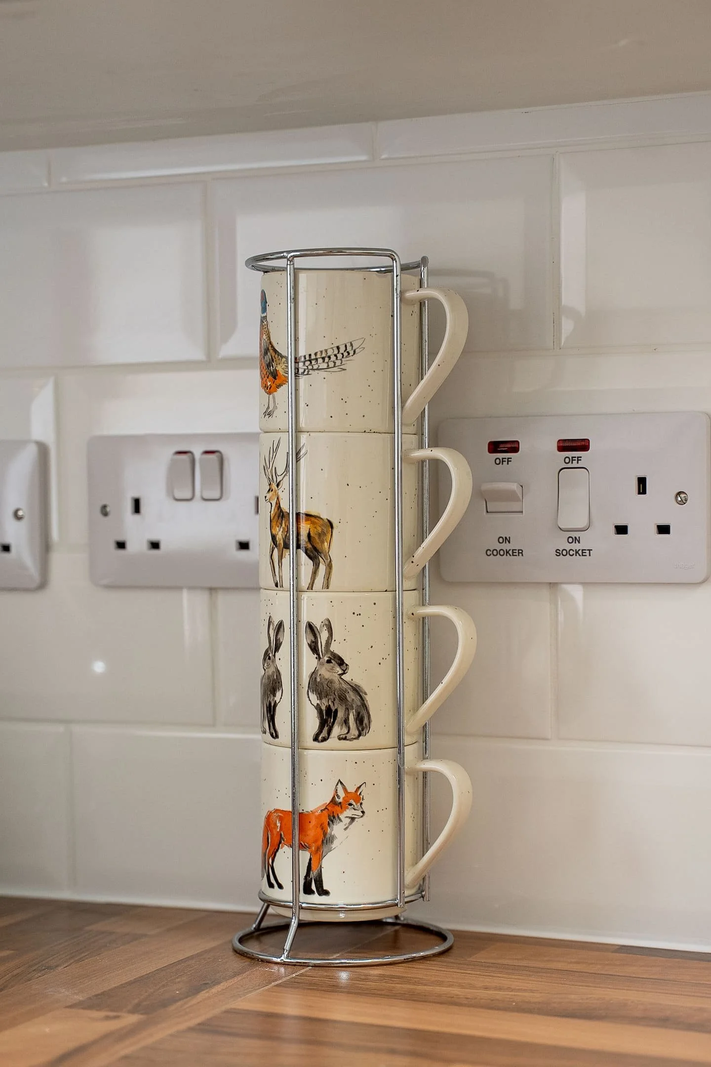 Four ceramic mugs with animal illustrations stacked on a metal mug rack on a kitchen counter, with electrical outlets and switches in the background.