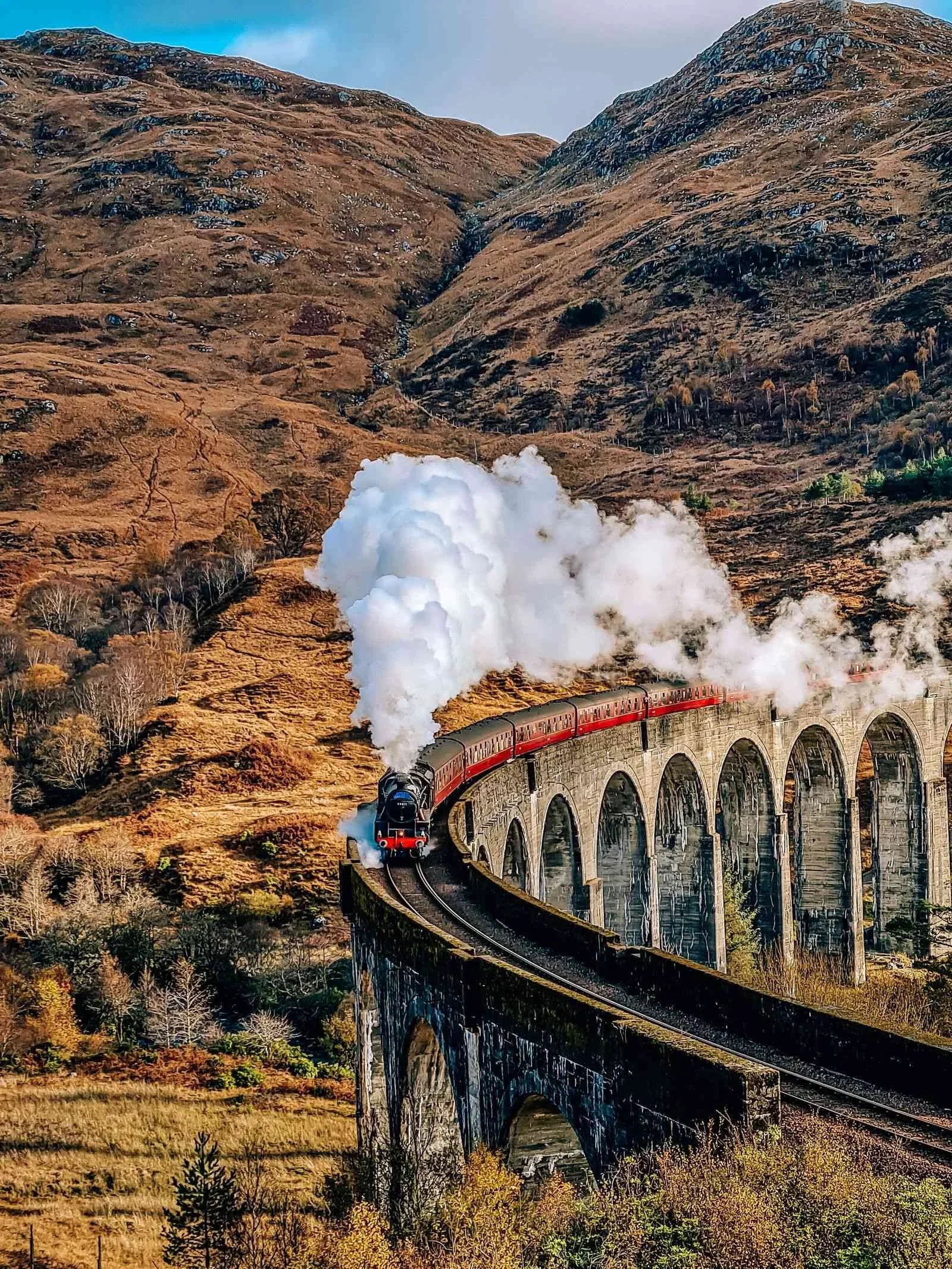 A vintage steam train with red cars crosses a large stone viaduct in a mountain landscape during autumn, with smoke billowing from the engine.