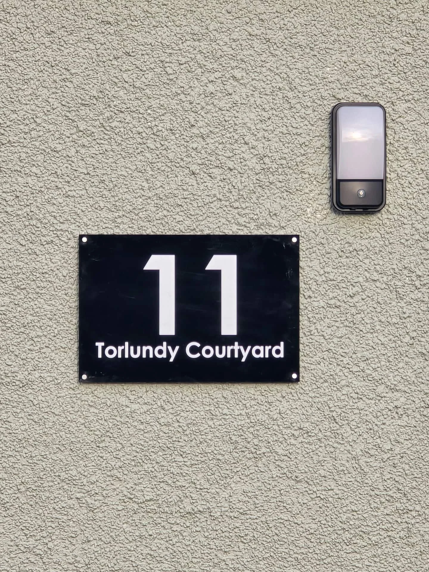 A black house number sign with the number 11 and the words 'Torlundy Courtyard' mounted on a beige stucco wall, with an outdoor light fixture above.