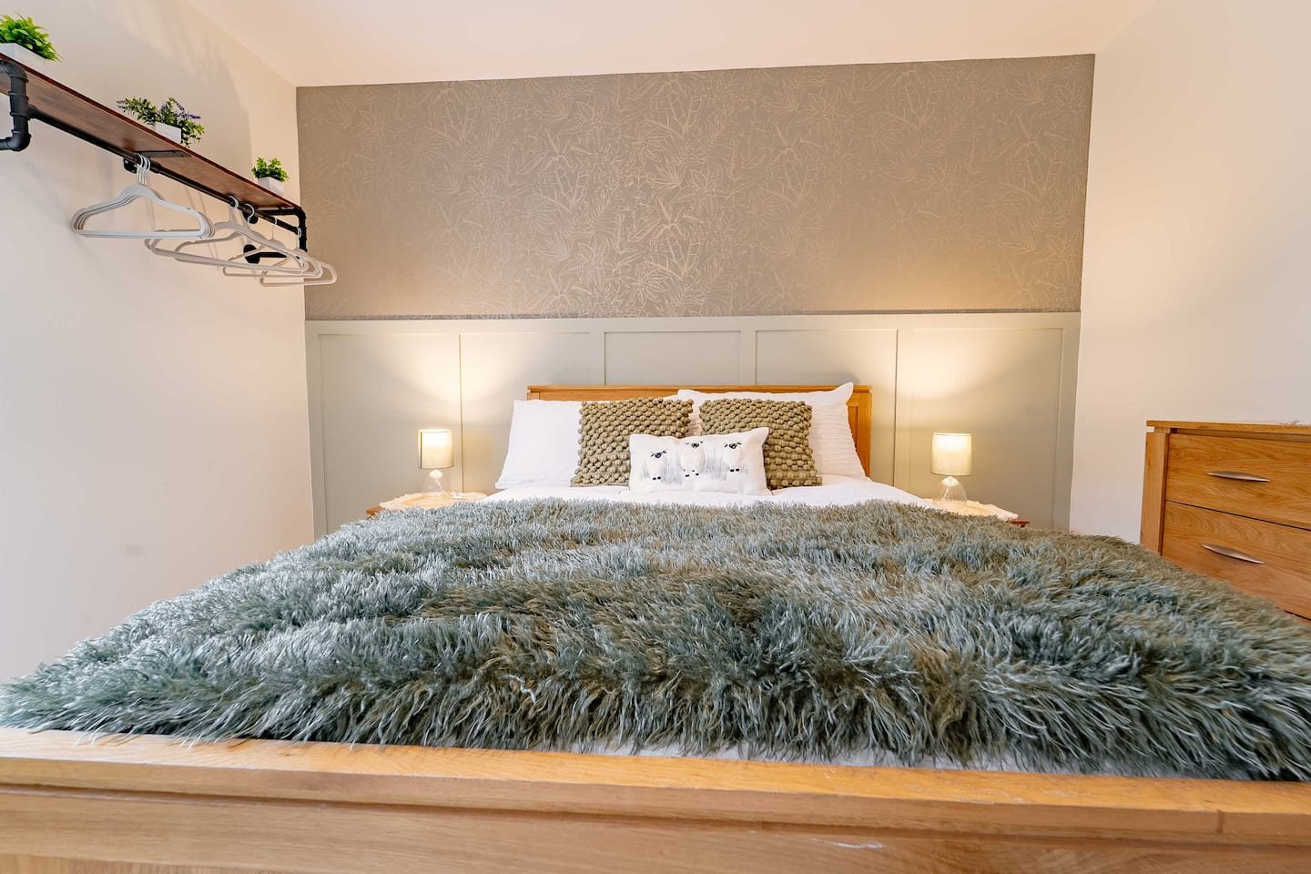 A neatly made bed with a fluffy gray blanket, decorative pillows, and two small bedside lamps in a cozy bedroom with beige walls and a wooden dresser.