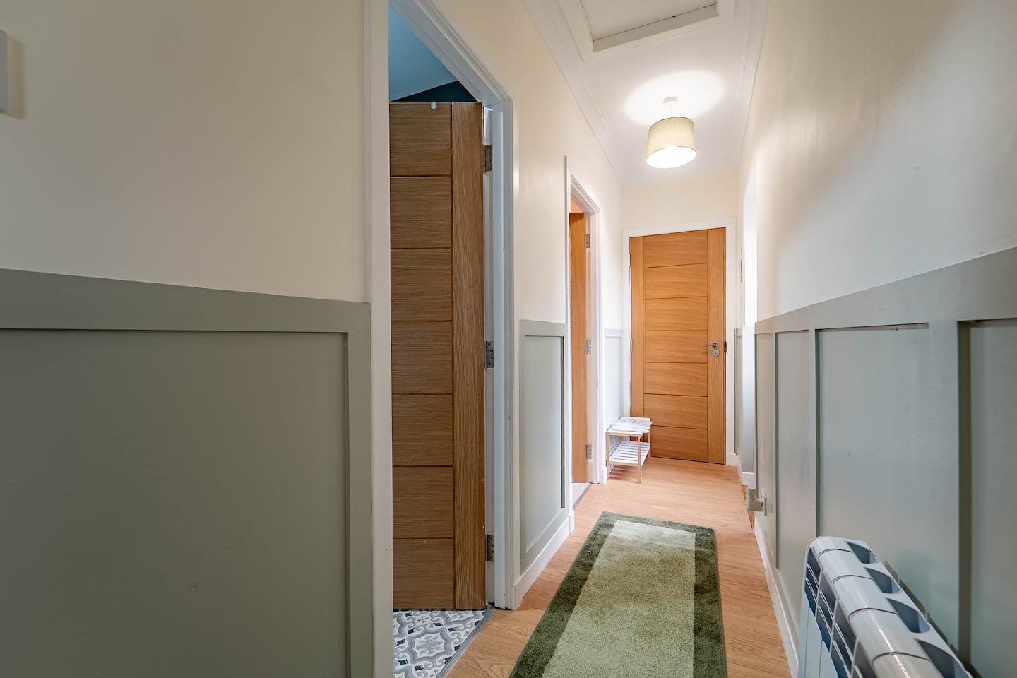 A hallway with light-colored walls and a wood floor, featuring a green runner rug, a wooden door at the end, and a white and blue heater on the right side.