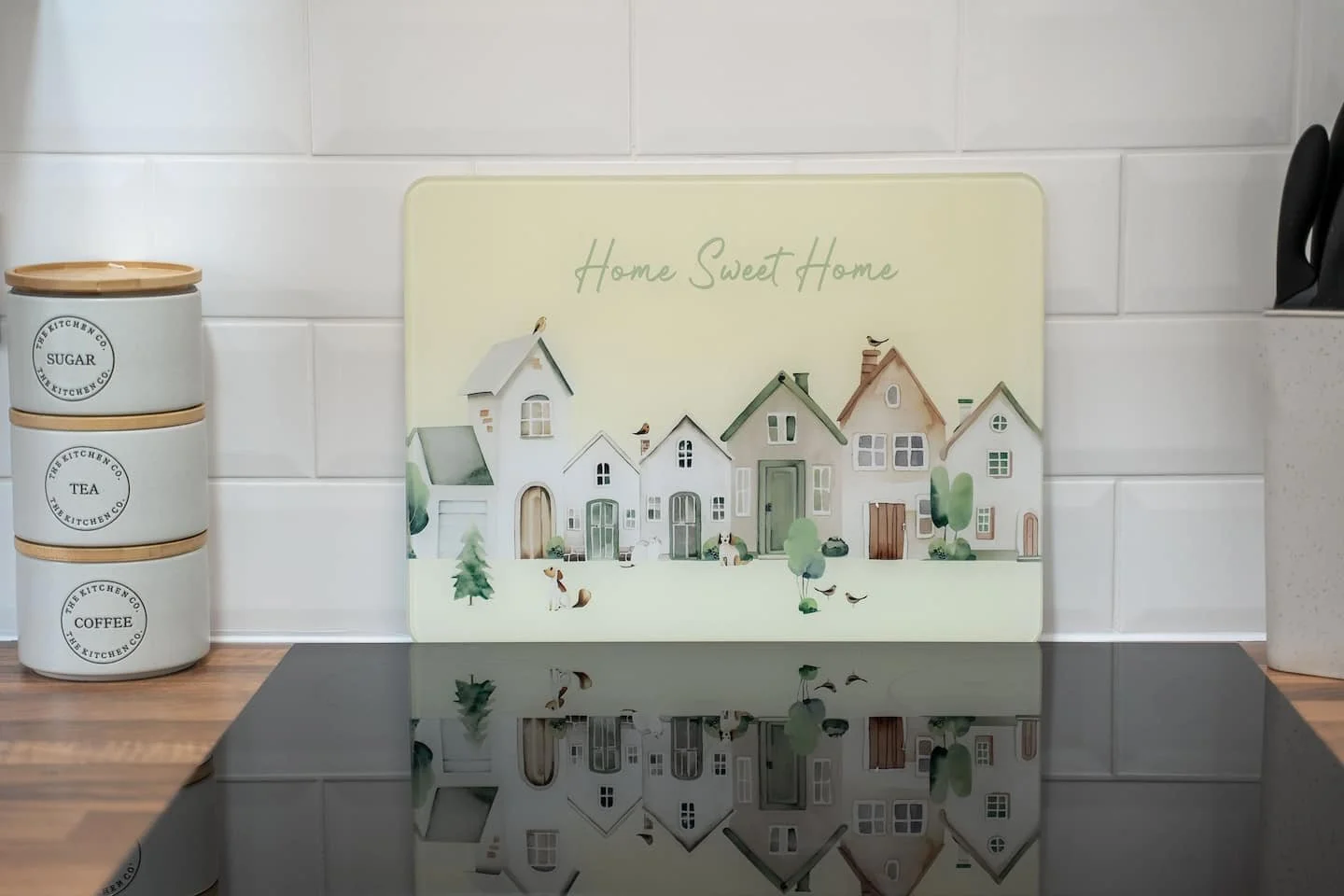 Kitchen countertop with a decorative sign saying "Home Sweet Home" featuring illustrated houses, a container labeled "Sugar," stacking containers for "Tea" and "Coffee," and a pair of black scissors.