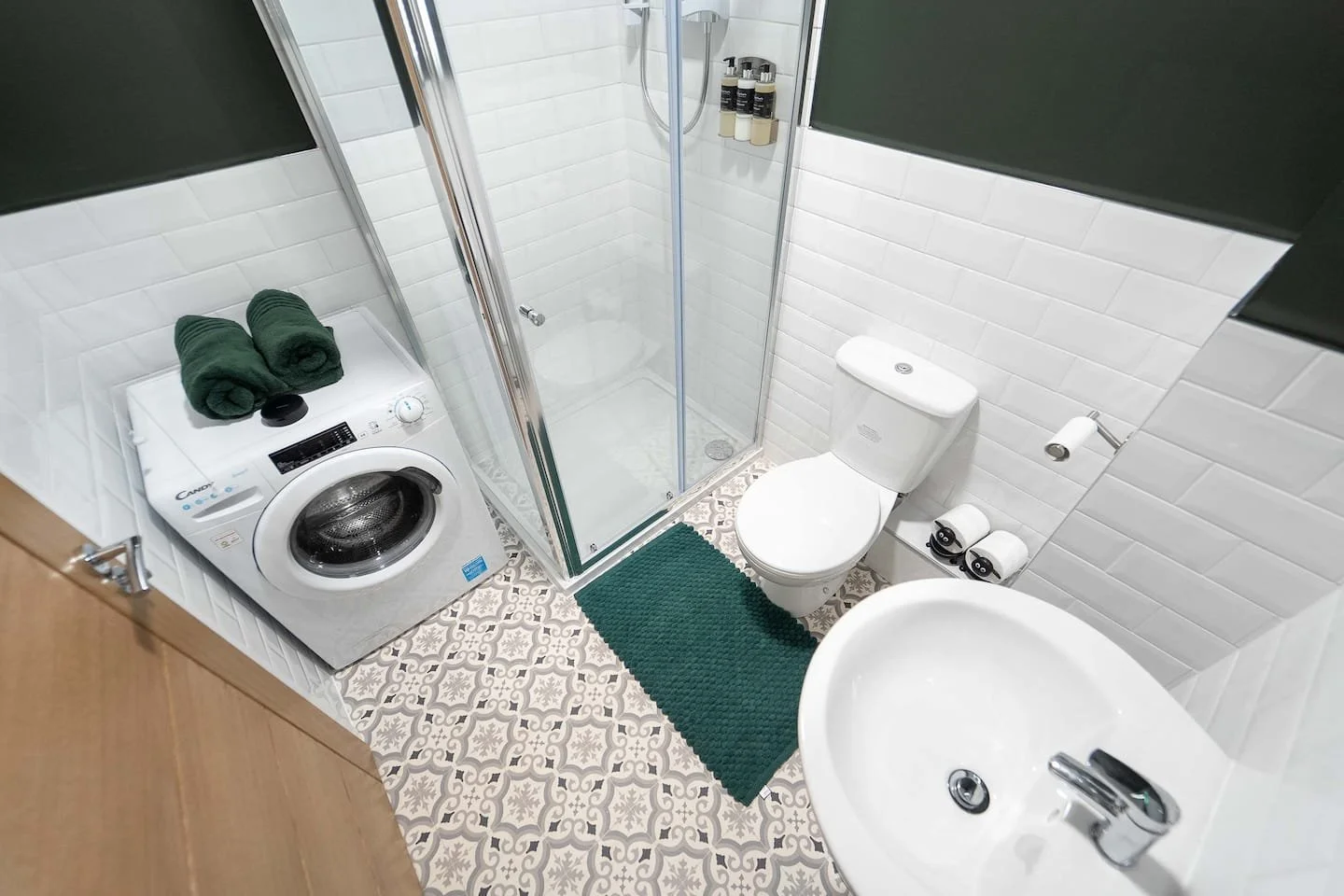 Small bathroom with patterned floor tiles, white tiled walls, a glass shower enclosure, a toilet, a small sink, a washing machine with two green towels on top, green accents including a rug and towels in the shower, and toilet paper rolls.