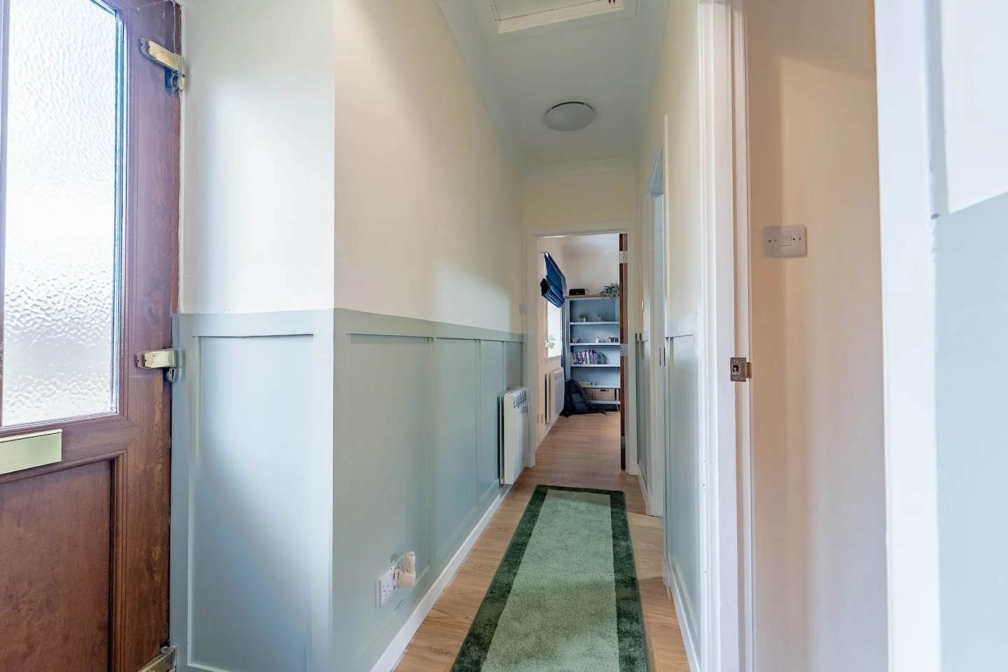 Bright hallway with wooden door, pale green wainscoting, and a green runner rug leading to a room with bookshelves and a window.