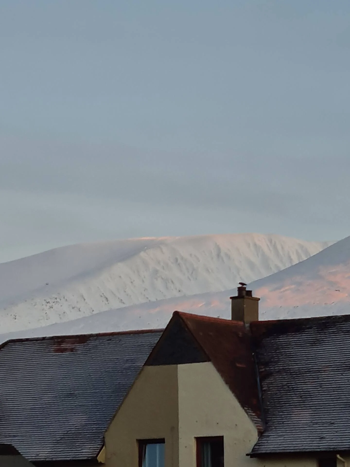 A house with a sloped roof covered in snow, with mountains in the background under a cloudy sky.