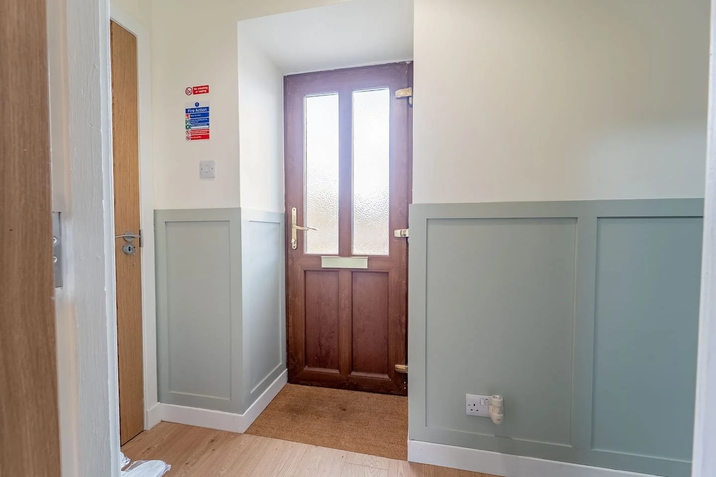 Interior view of a hallway with a wooden door and frosted glass window panels, light-colored walls with wall paneling, and a small brown rug on a wooden floor.