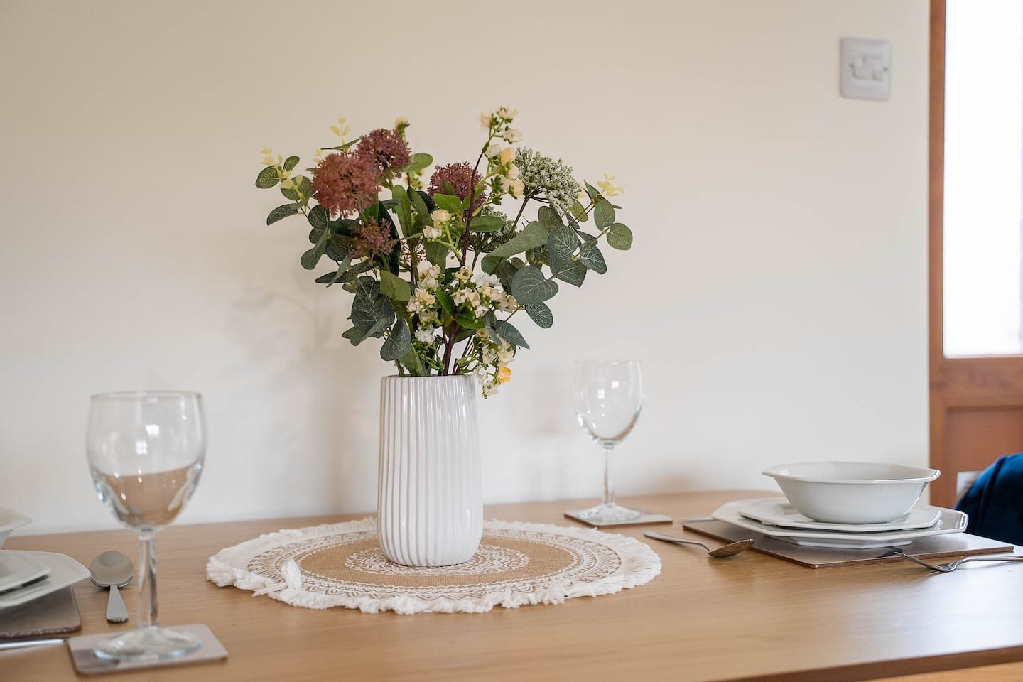 Dining table with white arranged dishes, wine glasses, and a white vase with mixed greenery and flowers on a lace doily.