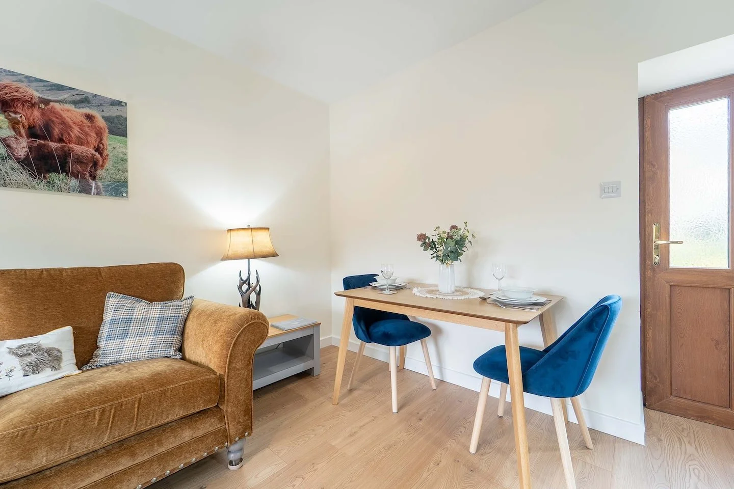 A living room with a brown sofa, a side table with a lamp, a wall art of a cow, a wooden dining table with two blue chairs, a vase with flowers, and a door with frosted glass