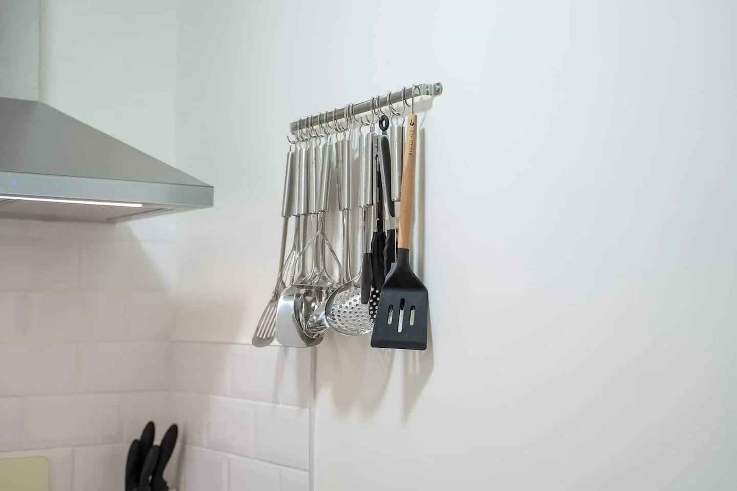 Kitchen wall with hanging utensils including a whisk, slotted spoon, ladle, spatula, and tongs, next to a stainless steel range hood.