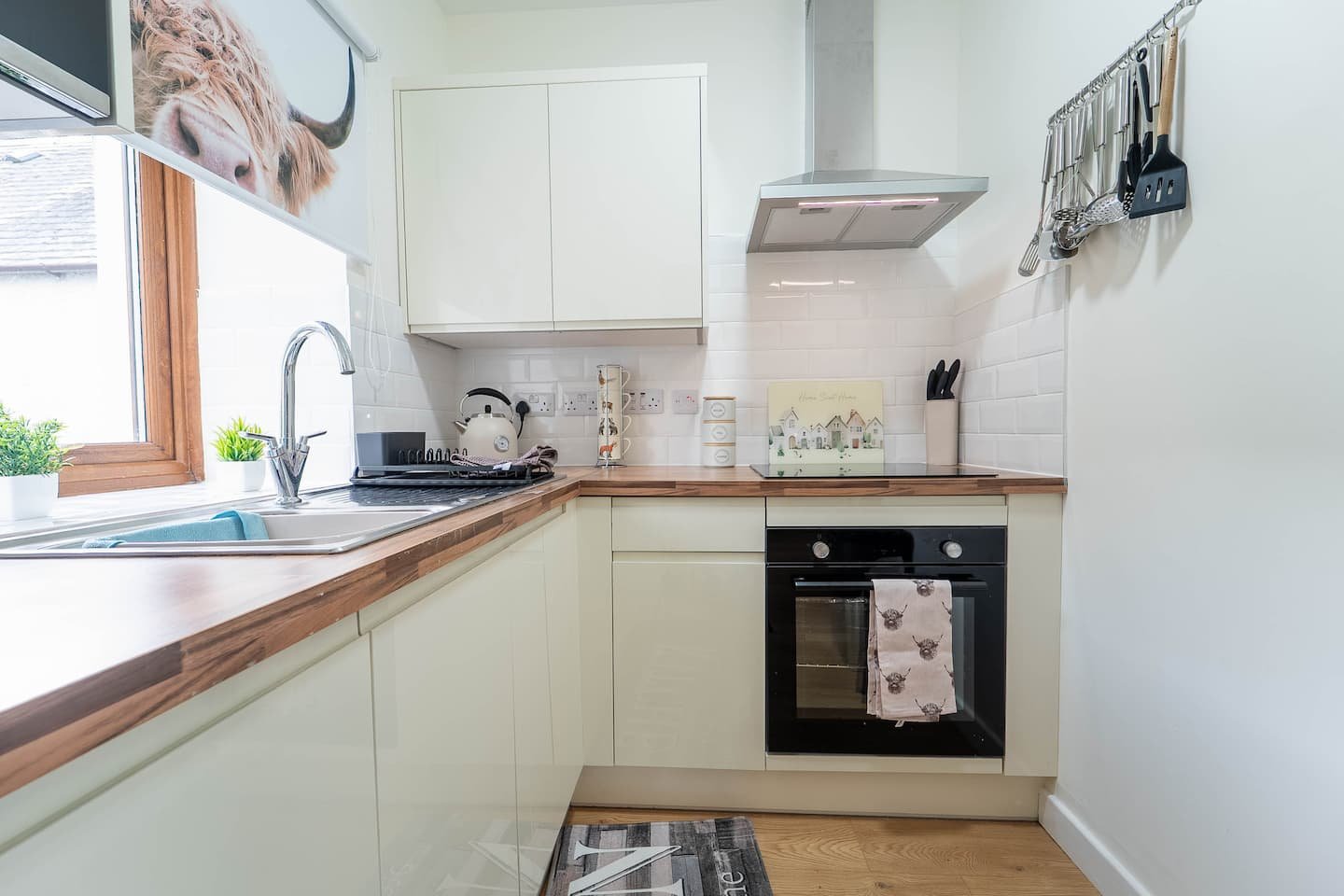 Clean kitchen with white cabinets, wooden countertop, and a window with a view outside. Contains a sink, stove, oven, knife holder, and hanging utensils on the wall.