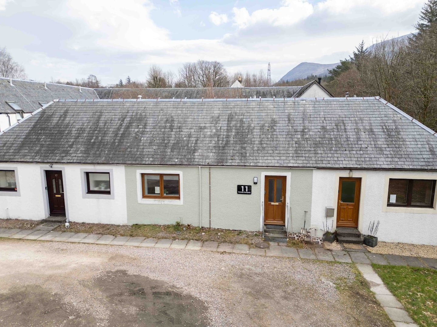 Row of single-story houses with gray tiled roofs, white and pale green walls, wooden doors, and small front steps, set in a rural area with mountains and trees in the background.