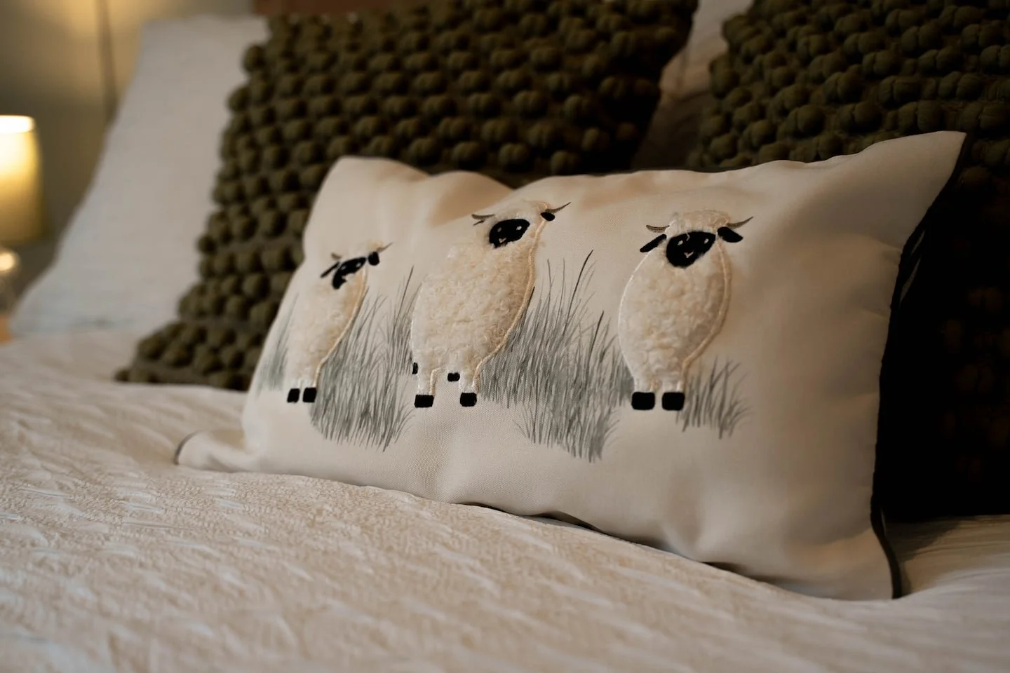 Decorative pillow with three cartoon-style sheep embroidery on a bed with textured white bedspread and dark woven headboard.