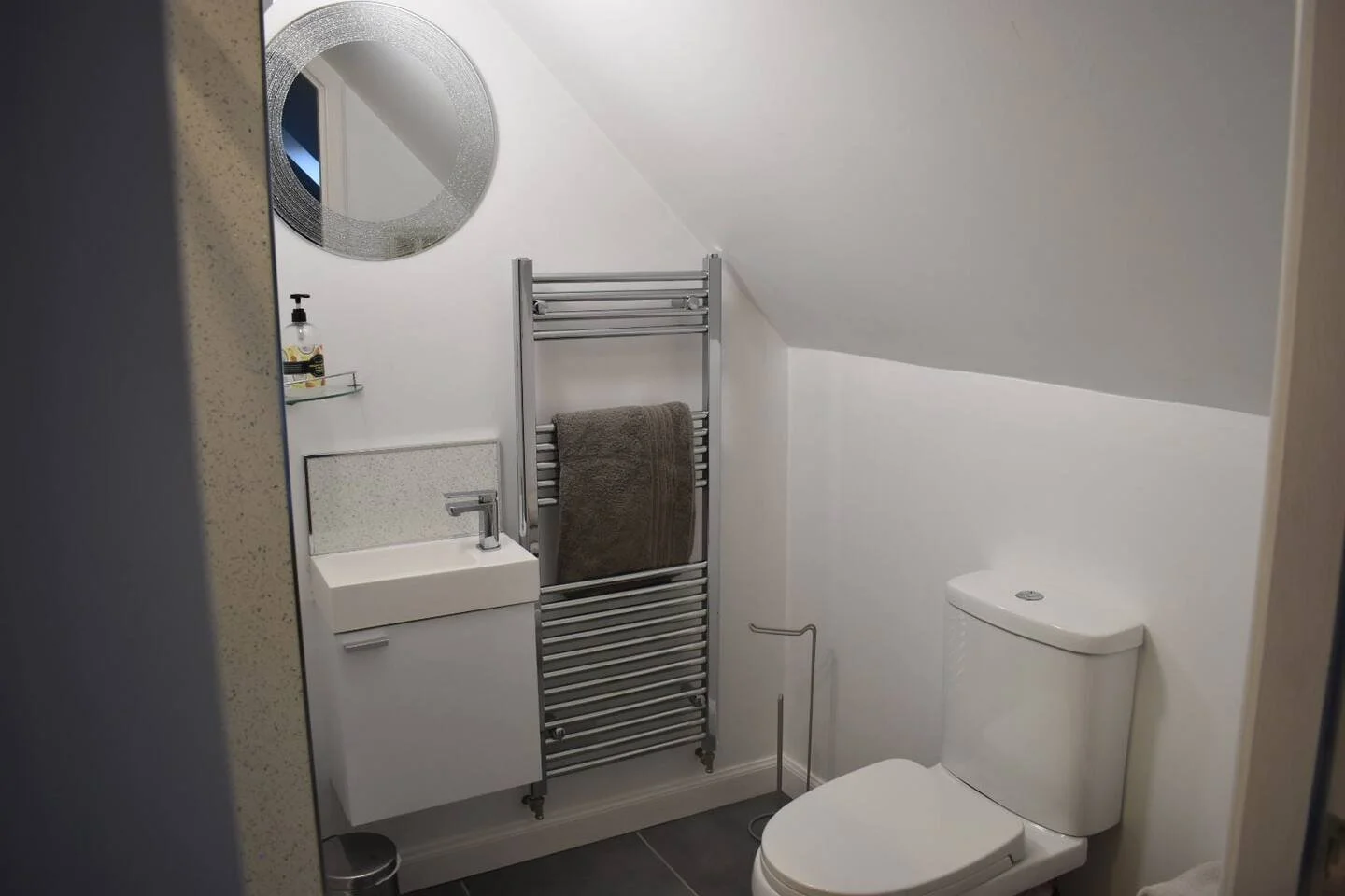 A small modern bathroom with a sloped ceiling, featuring a round decorative mirror, a small rectangular sink with a cabinet, a towel rack with a brown towel, a wall-mounted soap dispenser, and a white toilet.
