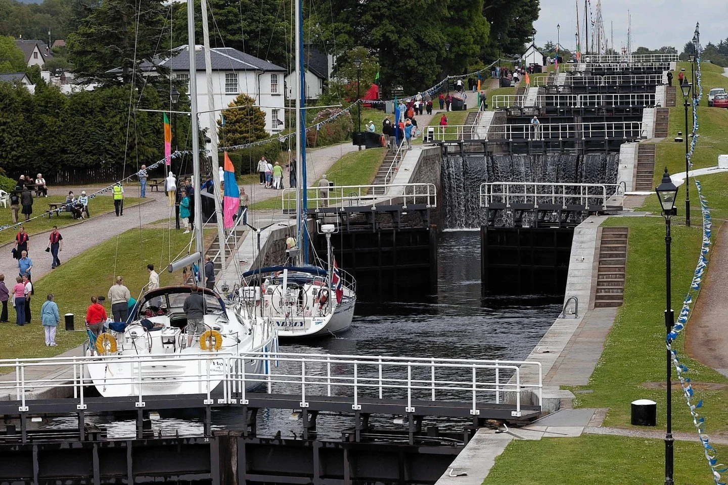 Two sailboats docked at a canal lock, with people walking and gathering on grassy slopes and pathways around the lock, as a row of houses and trees are seen in the background.