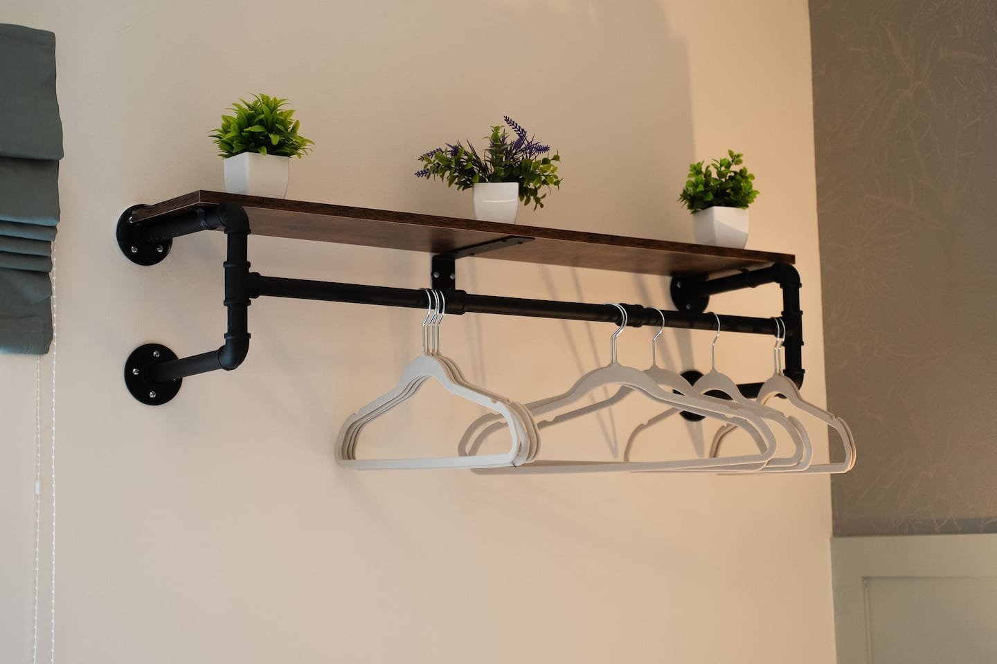 Wall-mounted clothing rack made of black pipe with a wooden shelf on top and three white hangers hanging below, decorated with three small potted plants on the shelf.