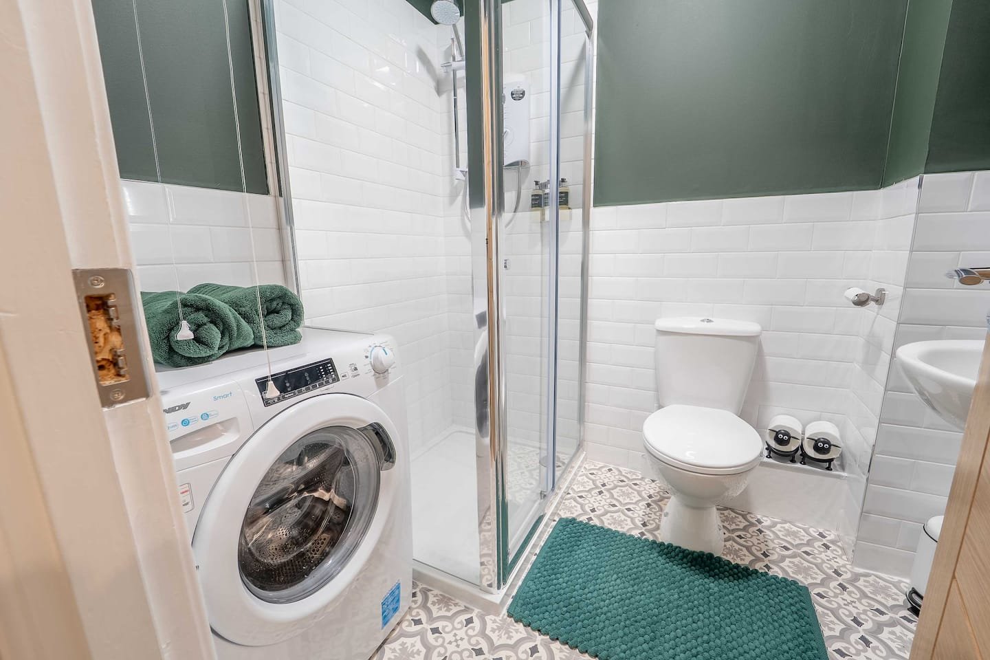 Bathroom with white tiled walls, green cabinet, washing machine, shower enclosure, toilet, small sink, and patterned tiled floor. Two green towels are folded on top of the washing machine.