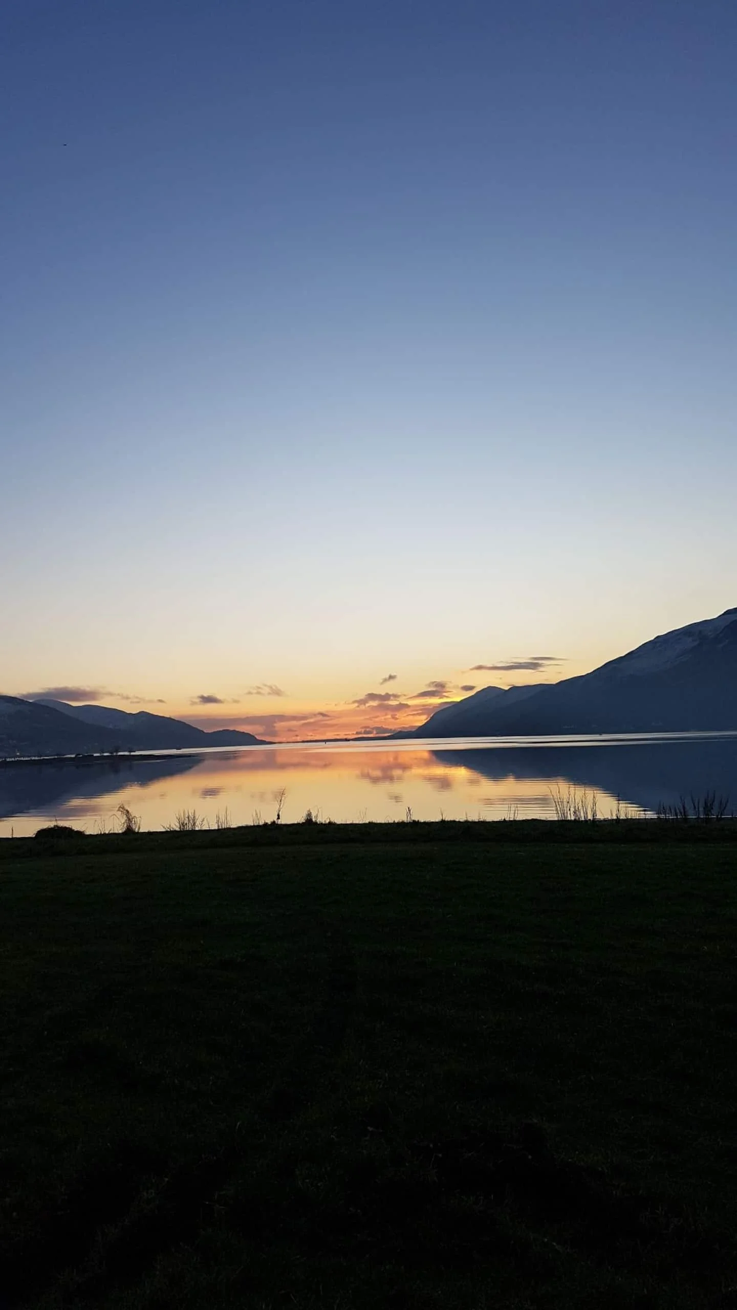 Sunset over a calm lake with mountains in the background and grass in the foreground.