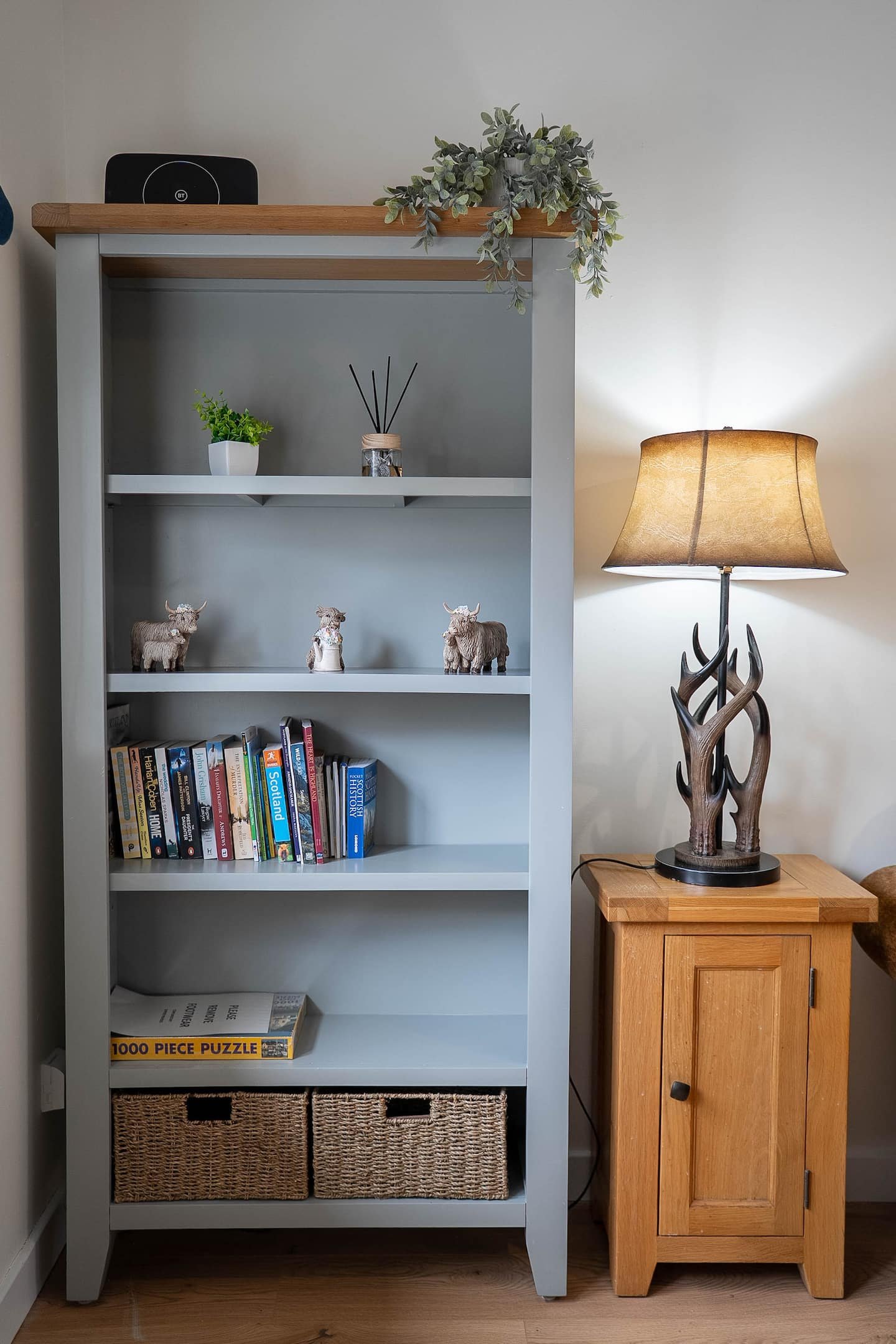 A tall gray bookshelf filled with books, toy moose, a potted plant, a diffuser, and a puzzle box, with a small wooden cabinet beside it. A lamp with a base made of antlers is on top of the cabinet, casting light in the room.
