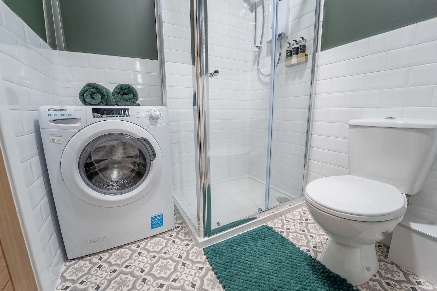 A small bathroom with a front-loading washing machine next to a glass shower enclosure and a toilet. Two rolled towels rest on top of the washing machine, and a teal rug lays on the patterned tile floor.