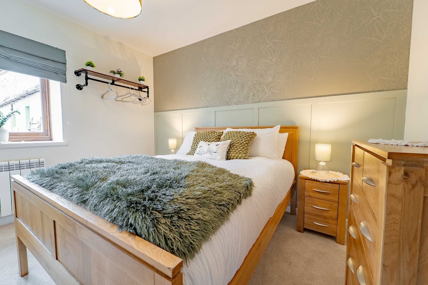 Cozy bedroom with wooden bed, plush gray blanket, white pillows, bedside lamps, wooden nightstand and dresser, window with a plant outside, and decorative wall.