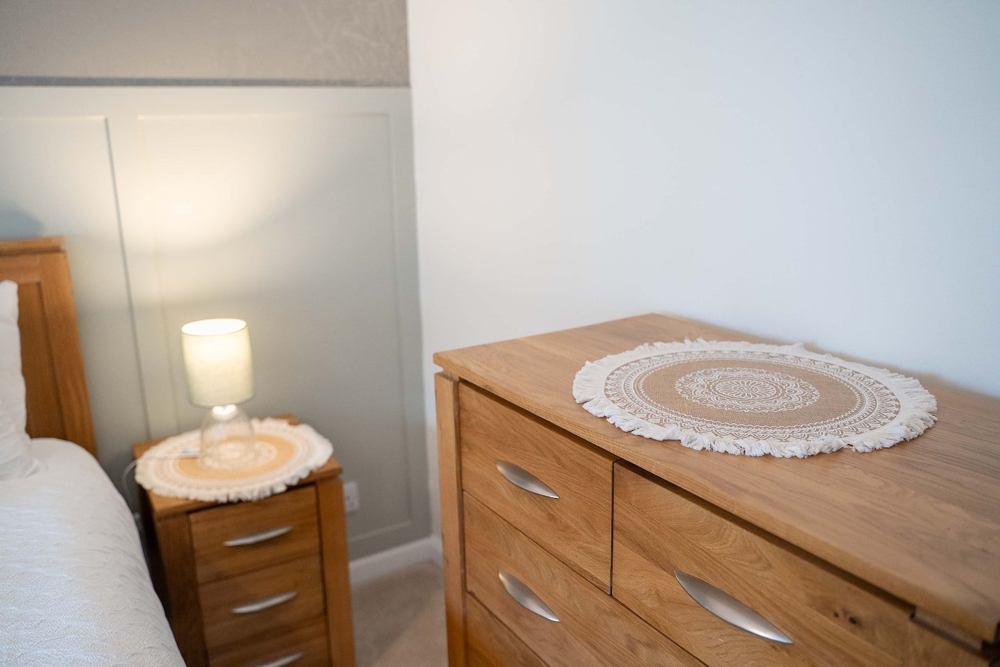 Bedroom with wooden furniture, a dresser with a round cloth on top, a bed with a similar cloth on the side table, and a lamp.